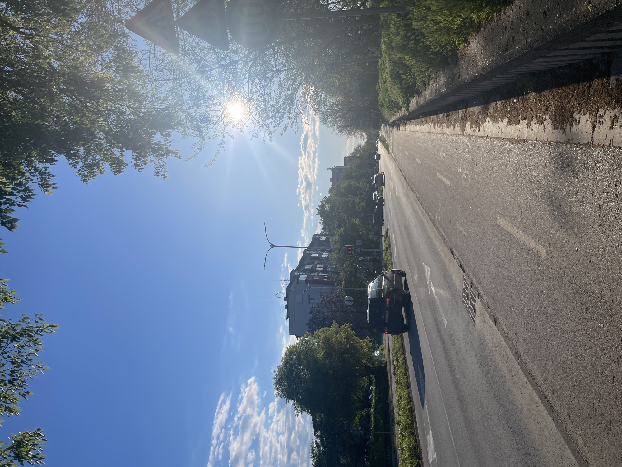 The image captures a sunny daytime view of a city street in Plovdiv, Bulgaria, oriented to depict the road stretching into the distance. The sun, partially obscured by green tree foliage, is bright in the upper left, casting a strong lens flare.
The main subject is an asphalt road with visible white lane markings, including a solid line separating the direction of traffic and a dashed line within a lane. A white arrow painted on the road indicates a forward direction. On the right side of the road, there is a paved sidewalk bordered by overgrown grass and small bushes. Several cars are visible on the road, with a dark-colored car in the foreground traveling away from the viewer. Its license plate, "PB 0975 CX," confirms the location as Plovdiv. Other vehicles are further down the street.
Along the left side of the road, dense green trees line the street. On the right, beyond the sidewalk and some greenery, multi-story residential or commercial buildings are visible, typical of an urban setting. The sky is clear blue with several scattered white cumulus clouds, stretching across the upper and central parts of the frame.
Notable details include several street signs. Prominently in the upper left, a white circular speed limit sign with a red border displays "30," indicating a speed limit of 30 km/h. Below it, a smaller rectangular sign with a red background shows "14.4," possibly a temperature reading or other information. A square drain cover is visible on the sidewalk to the right. The overall atmosphere is calm and bright, suggesting pleasant weather. No people are visible in the scene.