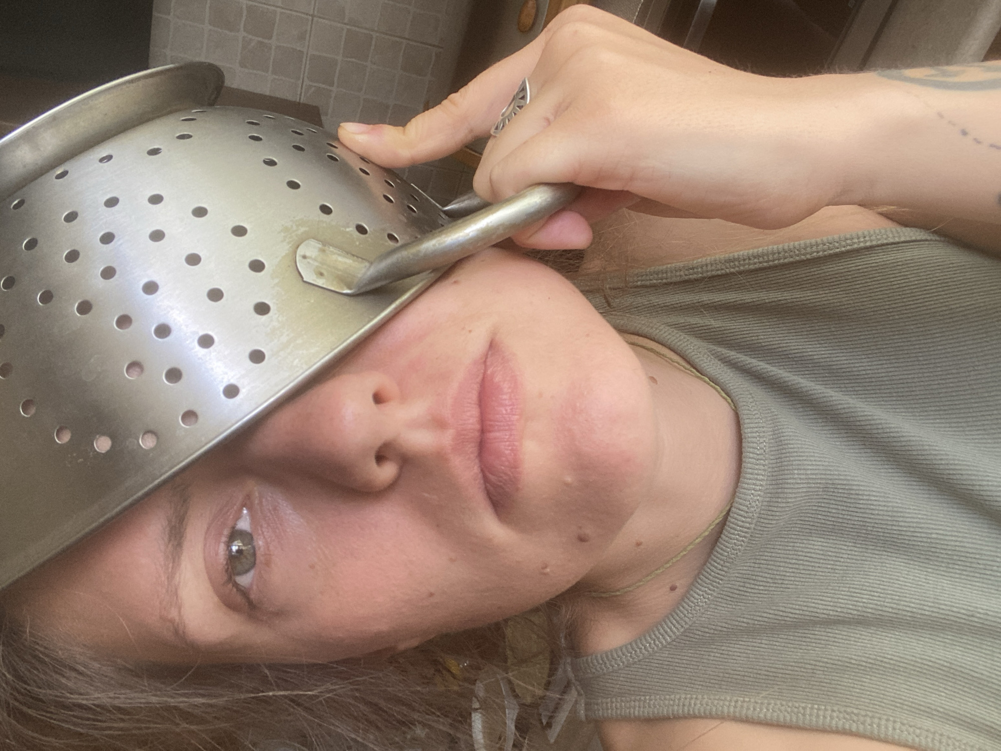 The image is a close-up, selfie-style shot of a woman's face and upper torso. A metal colander is held in front of the right side of her face, obscuring her eye and part of her nose and cheek. Her left eye is visible, with light brown irises and visible eyelashes. Her lips are slightly parted, and her skin appears fair with some freckles and minor blemishes. She is wearing a ribbed, olive-green tank top. A thin, light-colored necklace is visible around her neck. Her right hand is visible holding the colander, and a silver ring with a triangular design is on her ring finger. The background is blurred but appears to be a domestic environment, possibly a kitchen, with tiled surfaces visible. The lighting suggests daylight. The image does not provide any specific visual cues to determine its location in Almaty, Kazakhstan.