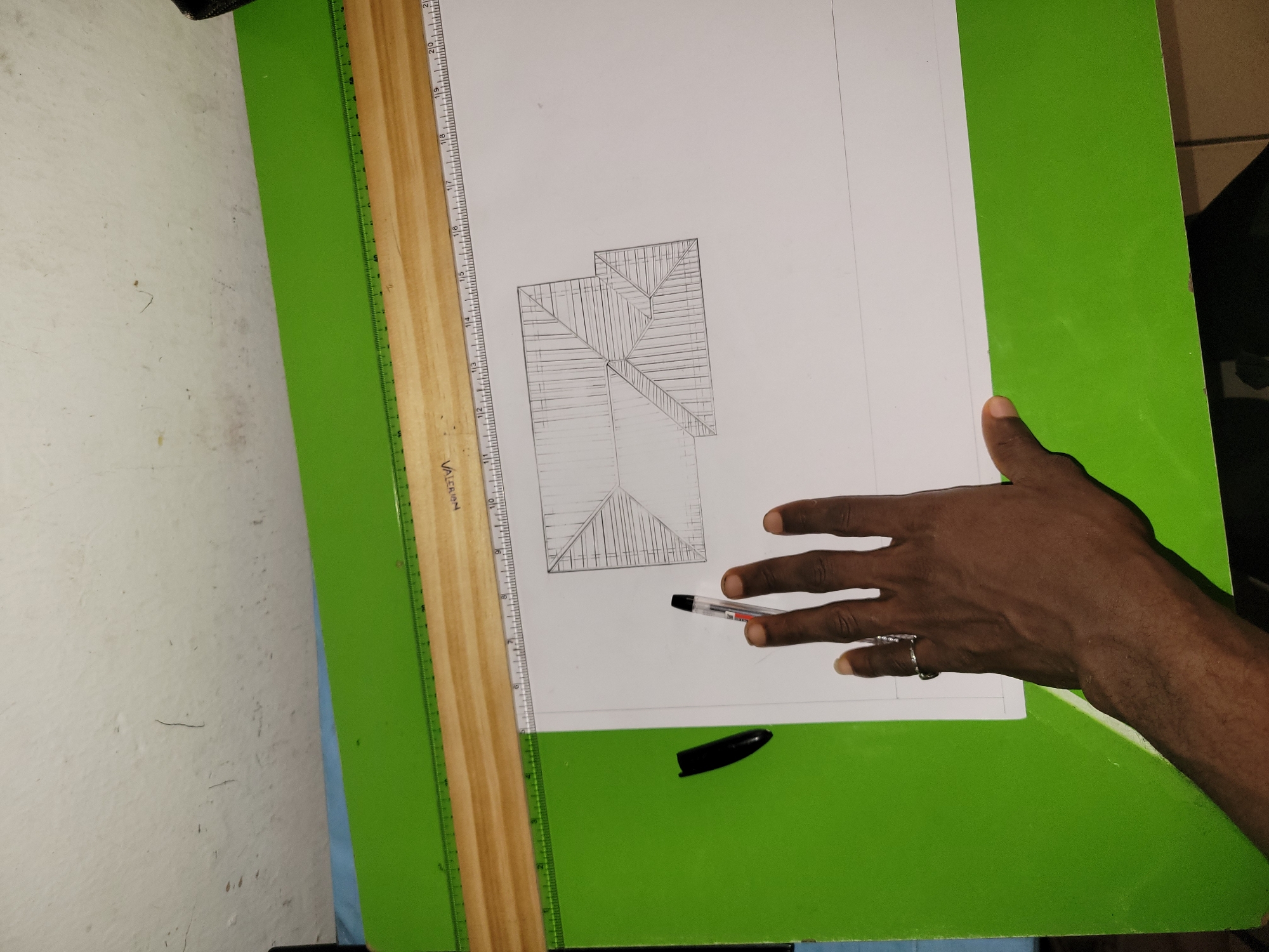 An overhead, eye-level shot shows a person's hand and arm extended across a drawing board, holding a pen. The hand is dark-skinned, with a ring on the index finger. The arm is also dark-skinned and partially visible. The person is drawing a detailed sketch of a house roof, with lines indicating rafters and roof tiles, on a white sheet of paper. The drawing board is green and is placed on a light wooden drafting table with a ruler attached. The background is a white, textured wall.