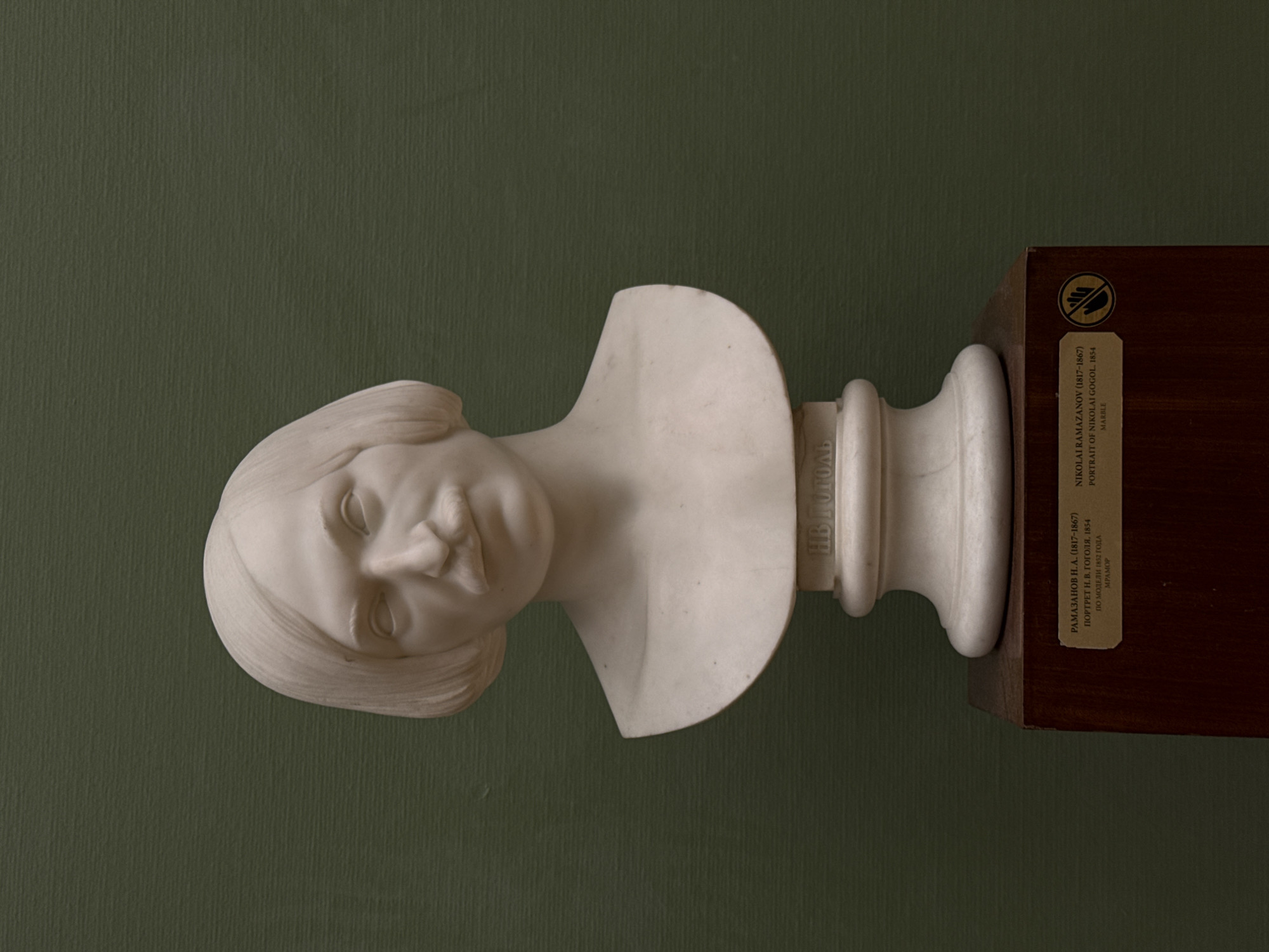 The image depicts a marble bust of Nikolai Gogol, sculpted in 1854 by Nikolai Ramazanov. The bust is displayed on a dark wooden stand against a green wall. The bust shows Gogol with a slightly tilted head and closed eyes. Inscription reads: "NIKOLAI RAMAZANOV (1817-1867) PORTRAIT OF NIKOLAI GOGOL. 1854 MARBLE" and "RAMAZANOV N. A. (1817-1867) PORTRET N. V. GOGOLYA, 1854 PO MODELI 1852 GODA MPAMOP" and name "HB GOGOL." The location is likely in Moscow, Russia, but the exact museum or gallery is not explicitly indicated in the image.