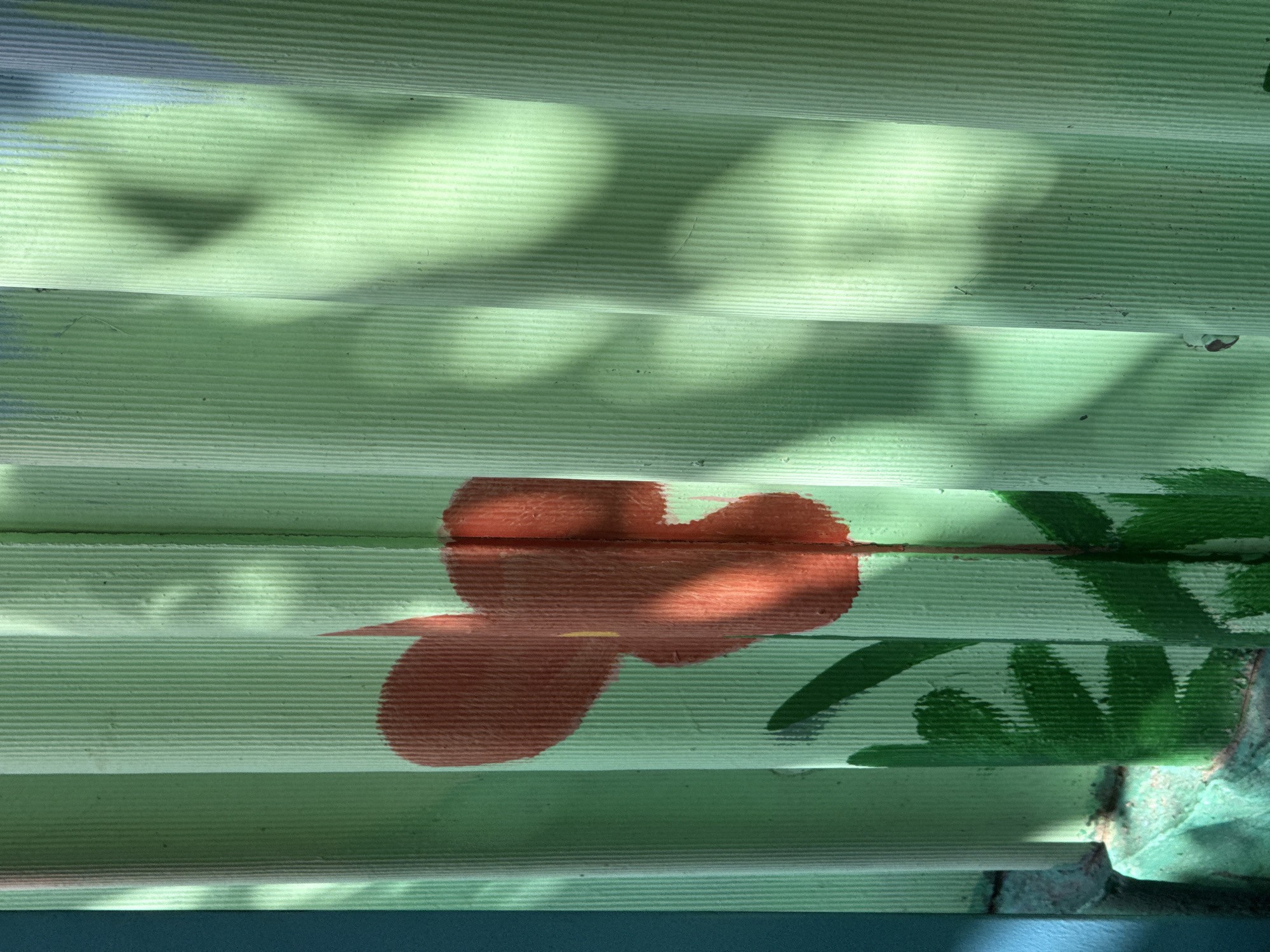 Here's a description of the image:
The image presents a close-up view of corrugated siding, painted a light green color. There is a hand-painted floral design in the center, with a red flower and green foliage. Portions of the image are covered in shadow.