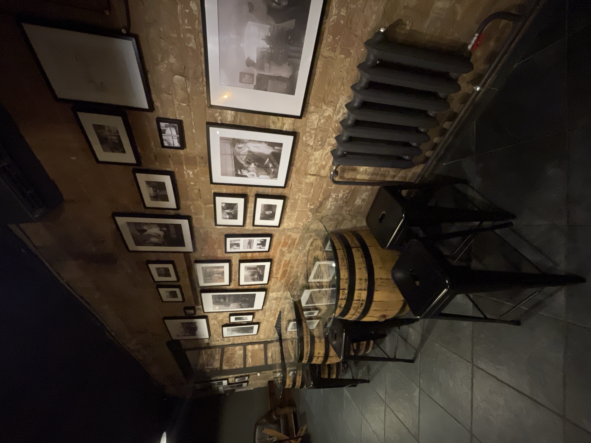 The image shows the interior of a bar or restaurant. The walls are brick with a collection of framed black and white photographs. There is a black radiator mounted on the wall. Several barrel tables with glass tops are accompanied by black chairs. The floor is tiled in a dark color. The location is likely Krasnoyarsk, Russia, based on the context. There are no people visible in the image. The scene appears staged and unoccupied.