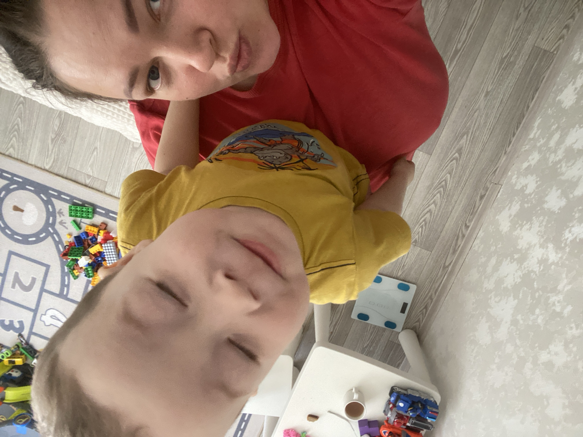 A selfie shows an adult female with dark hair and a red shirt, likely indoors. She is positioned above a young boy in a yellow shirt with a graphic on it. The boy's eyes are closed. In the background, there is a play mat with numbers, a collection of toy bricks, a scale, a white table, a cup, and more toys. The location is likely indoors, in Sochi, Russia.