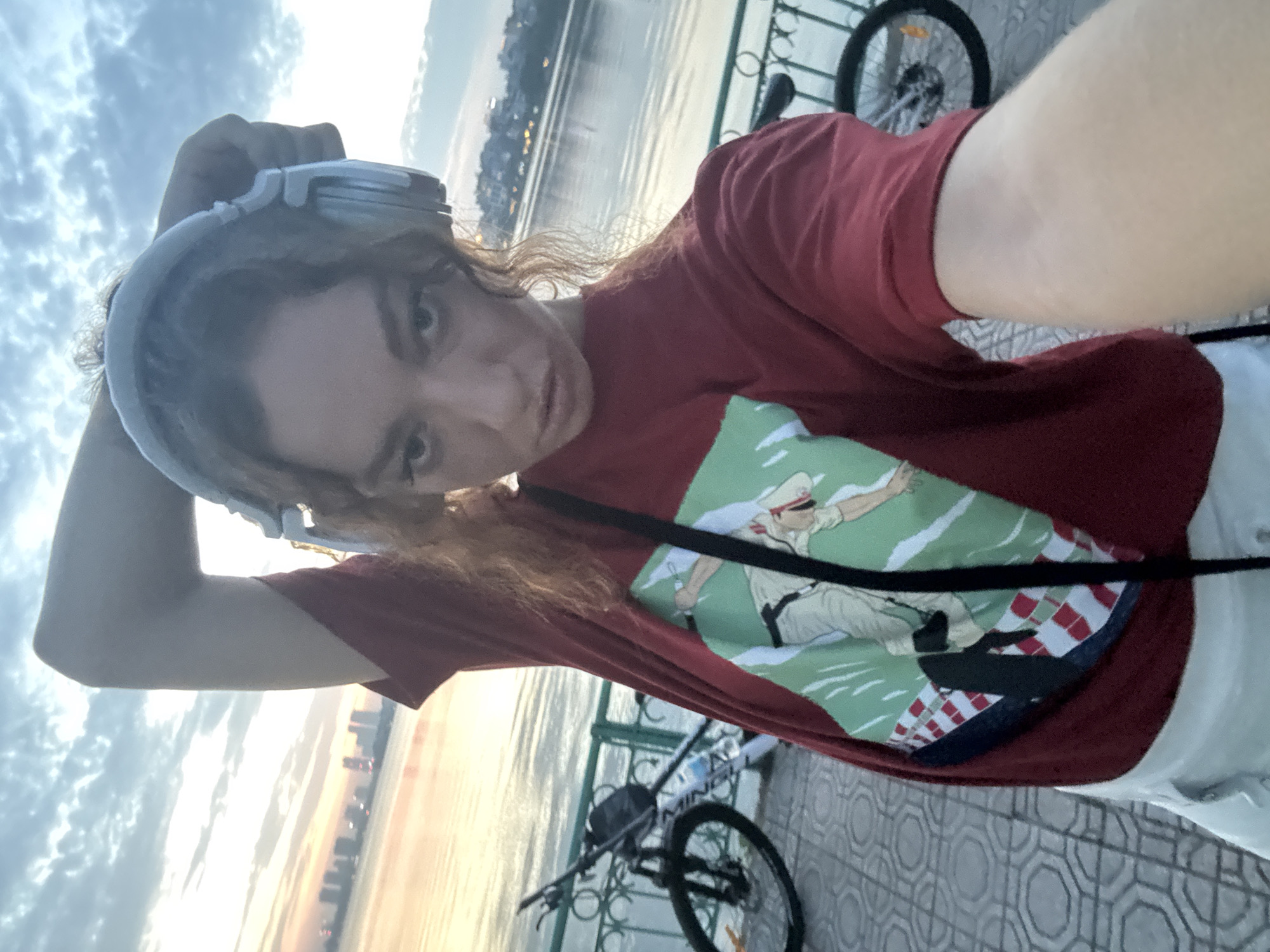 In Hanoi, Vietnam, at sunset, a young woman stands on a paved walkway by a body of water, likely West Lake, with a city skyline visible in the distance. She is wearing a red t-shirt with a graphic print, white shorts, and headphones. Her left arm is raised behind her head, and she is looking at the camera, holding it to take a selfie. Two bicycles are parked behind her, one to her left and one to her right. A decorative metal railing runs along the edge of the walkway. The sky is filled with clouds, tinged with the warm colors of the setting sun.
