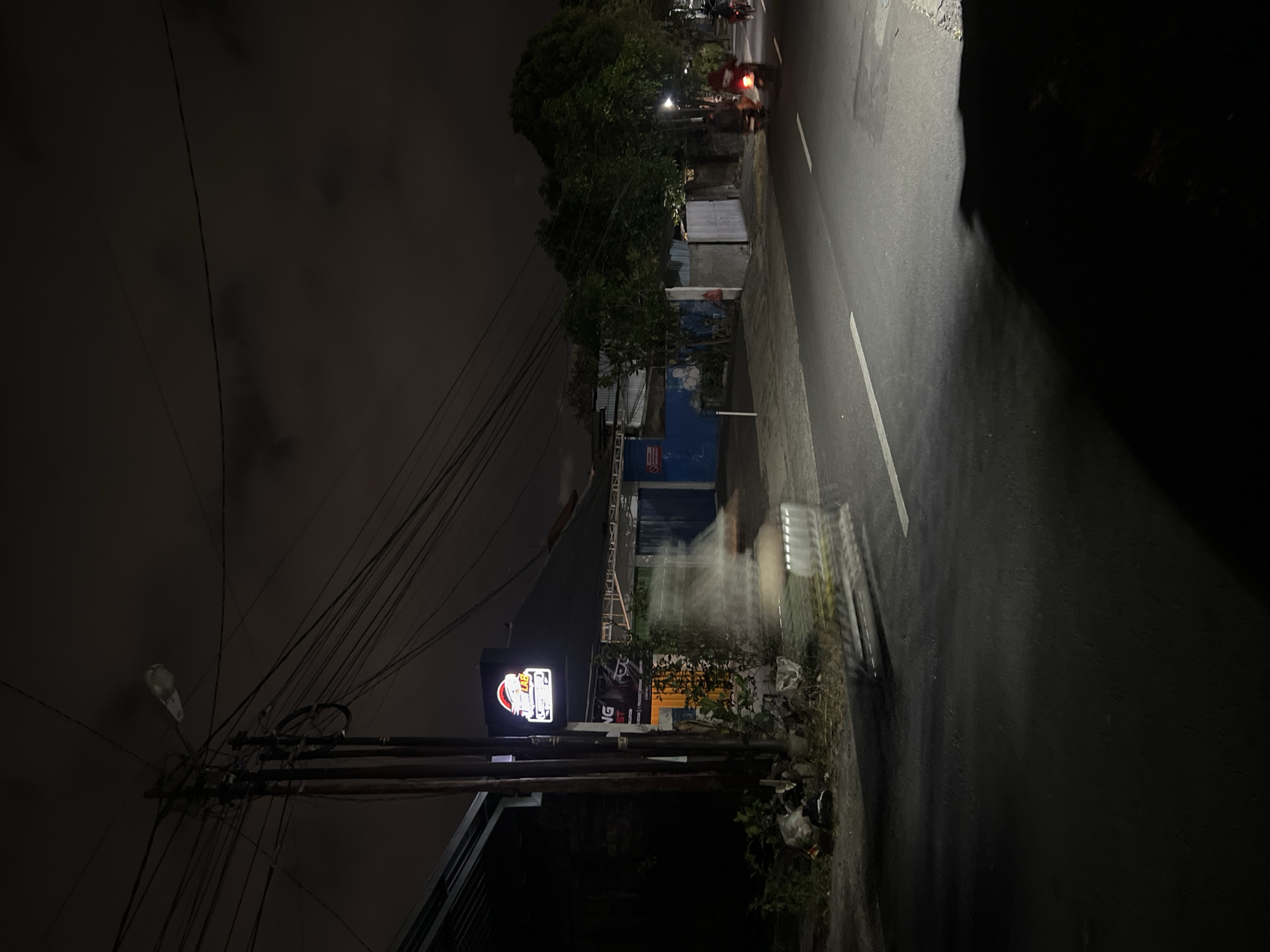 The image depicts a nighttime street scene in Denpasar, Indonesia. A dimly lit street runs diagonally across the frame, with a white line marking its center. On the left side of the street, a dark building with a glowing sign is visible. The sign is rectangular with rounded corners and features a cartoonish illustration of a helmet with what appears to be a face inside, along with text that is partially obscured but includes the letters "NG ST". Adjacent to the building is a utility pole with numerous wires crisscrossing overhead. In front of the building, a blurry, elongated figure, possibly a person or a vehicle in motion, is captured in the foreground, creating a streaking effect due to the long exposure. To the right of the street, trees and vegetation line the roadside, and further down the street, several motorcycles with illuminated headlights and taillights are present, with one motorcycle clearly visible in motion with a rider. The sky is dark and cloudy, typical of nighttime in a tropical environment. The overall atmosphere is one of a quiet, urban street after dark.