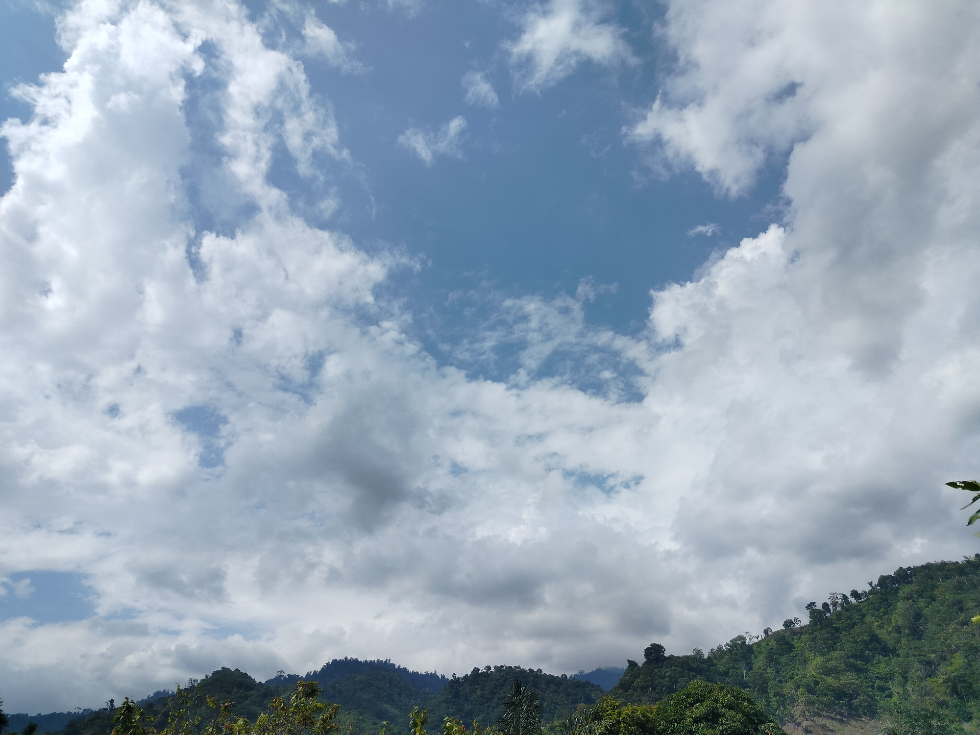 The image depicts a landscape dominated by a sky filled with a mix of white and grey clouds. The lower portion of the image shows rolling hills covered in dense green vegetation. The terrain appears to be rural and possibly mountainous. There are no visible people or man-made structures in the image, suggesting a natural or remote setting. The scene is consistent with the geography of Tegarmiko, Indonesia, which is known for its lush, hilly terrain and tropical climate, often characterized by such cloud formations.