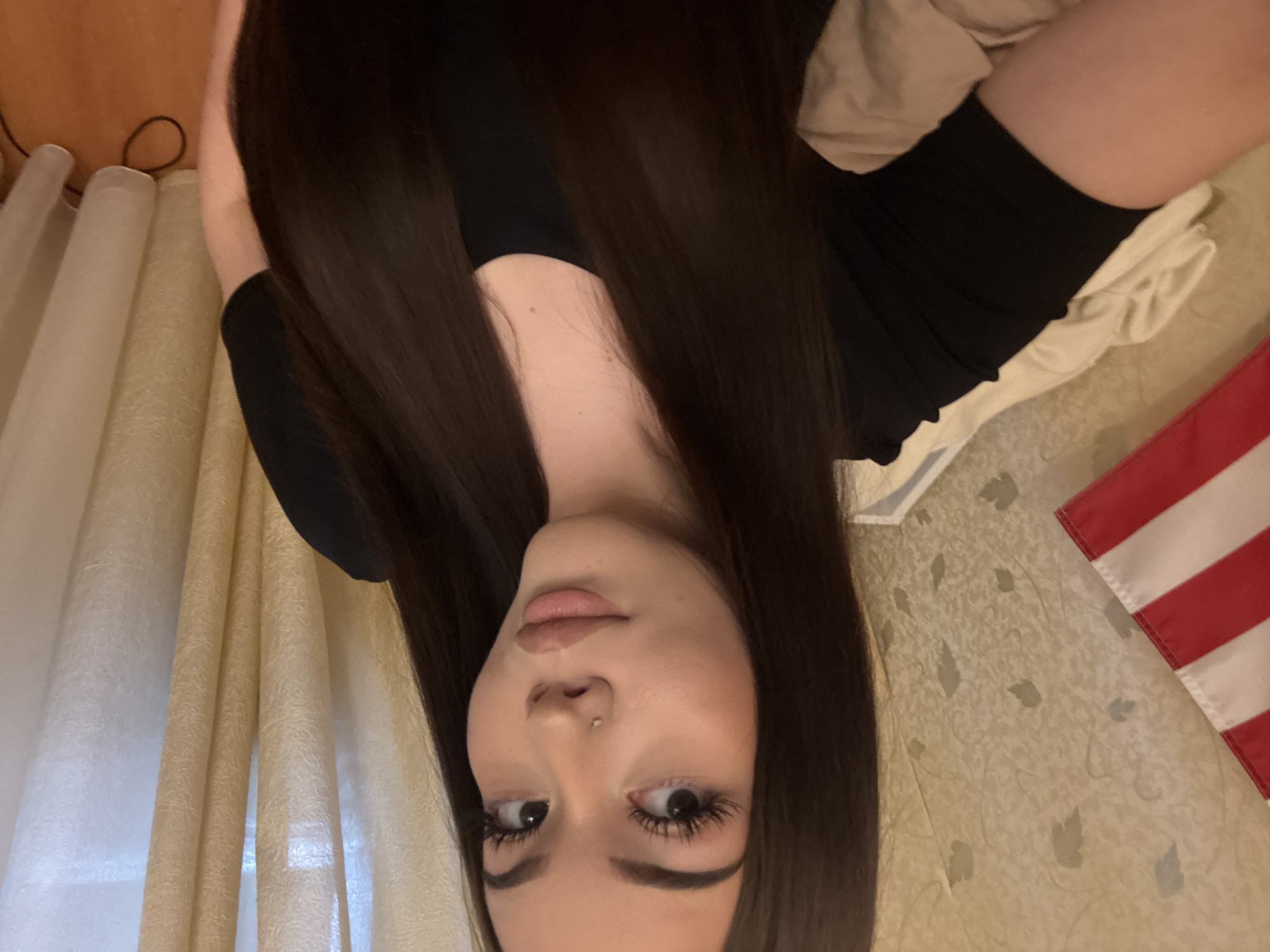 The image, taken in Naberezhnye Chelny, Russia, is oriented 90 degrees clockwise and features a young woman lying down indoors. She has long, straight, dark brown hair that frames her face and drapes over her shoulders. She is wearing a fitted black long-sleeved top. Her face shows a neutral expression, with her gaze directed towards the camera. Her lips are full and slightly parted, and she has a small silver stud piercing on her right nostril. Her eyes are accentuated with dark eyeliner and mascara, giving the appearance of long eyelashes.
The background suggests an indoor residential setting. To the left side of the frame (when correctly oriented), light-colored, textured curtains are visible, appearing translucent. To the right, a wall is covered with a light, patterned wallpaper featuring subtle leaf designs. In the lower right corner, a portion of a red and white striped flag is visible, possibly decorative. The woman is resting on a light, textured surface, likely a bed or couch, with a light-colored pillow or blanket visible near her head. The lighting is ambient, and there is no visible text.