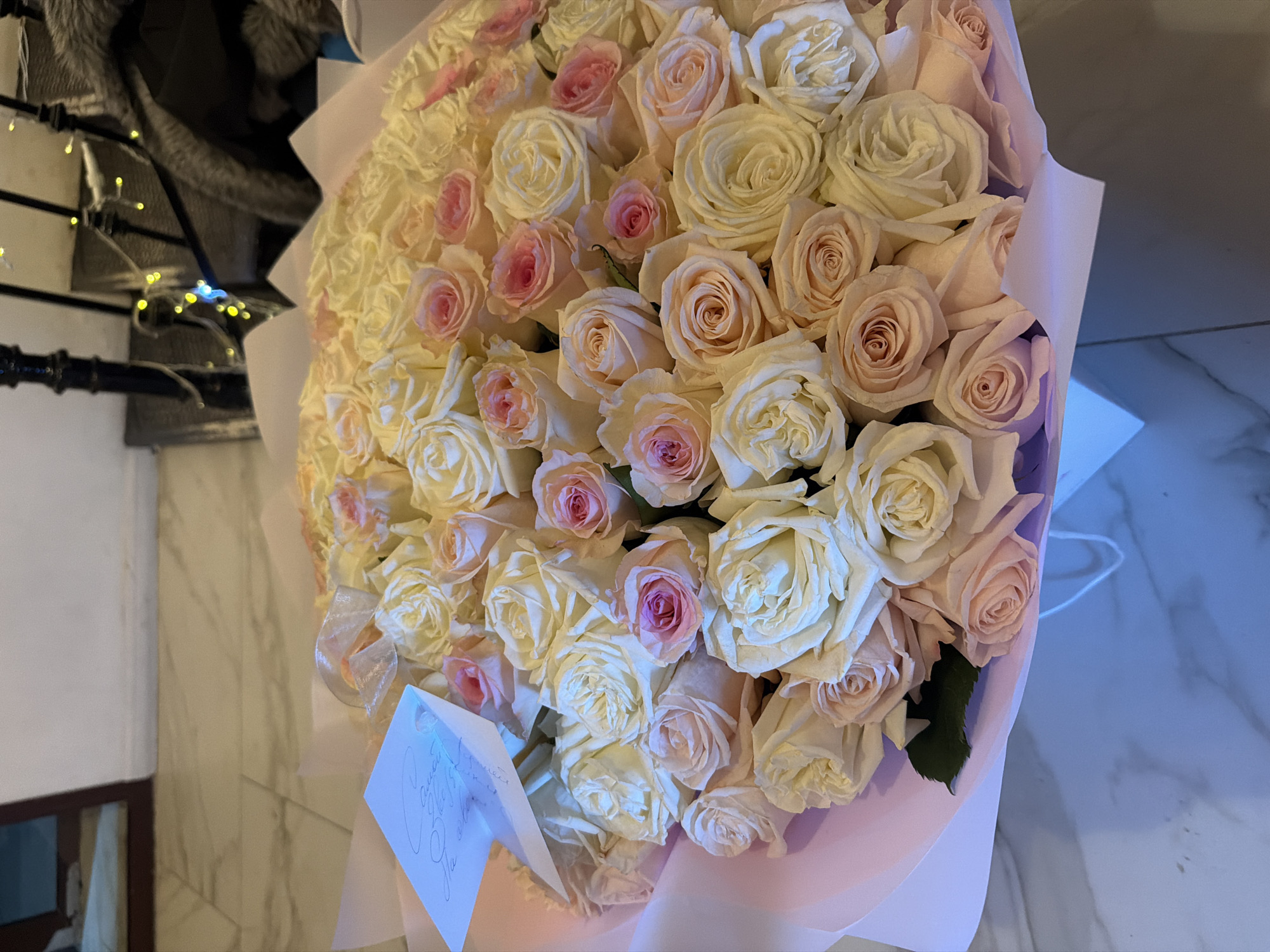 A bouquet of light pink and white roses is prominently featured. The flowers are arranged densely, wrapped in pale pink paper. A small card is visible near the bottom left corner. The scene includes a glimpse of a tiled floor with marble-like veins. Part of a window frame and what appear to be decorative lights are in the background, slightly out of focus. Based on the style of the scene, including the floor tiles and overall aesthetics, it may be a modern or recently renovated interior. While it is not possible to confirm the location of Moscow, Russia from the image alone, it is possible that this photograph was taken in a residential or commercial area within that city.