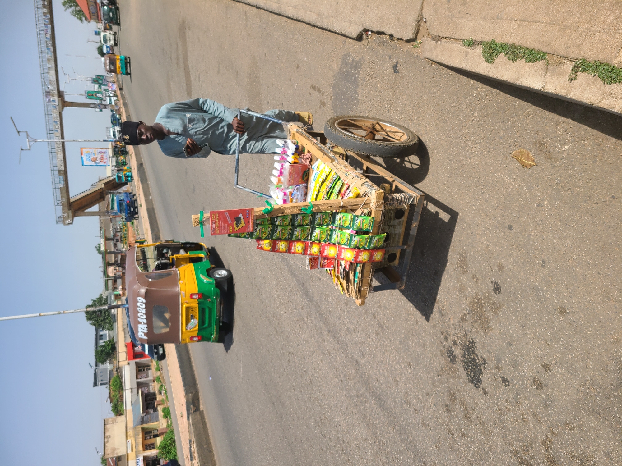 The image shows a street scene, probably in the city of Unknown, Unknown. A man wearing a light blue shirt, dark pants and a dark hat is pushing a wooden cart loaded with an assortment of items, possibly snacks, cigarettes and small household goods, along a paved road. The cart has a single bicycle-like wheel on one side.
To the left of the cart, a green and brown auto rickshaw with the license plate "PTA-10209" is driving on the same side of the road. In the background, there is a flyover, other vehicles, buildings and utility poles. A billboard can be seen on the flyover, partially obscured by infrastructure. The weather appears to be sunny with clear skies. The scene is captured from a higher vantage point.