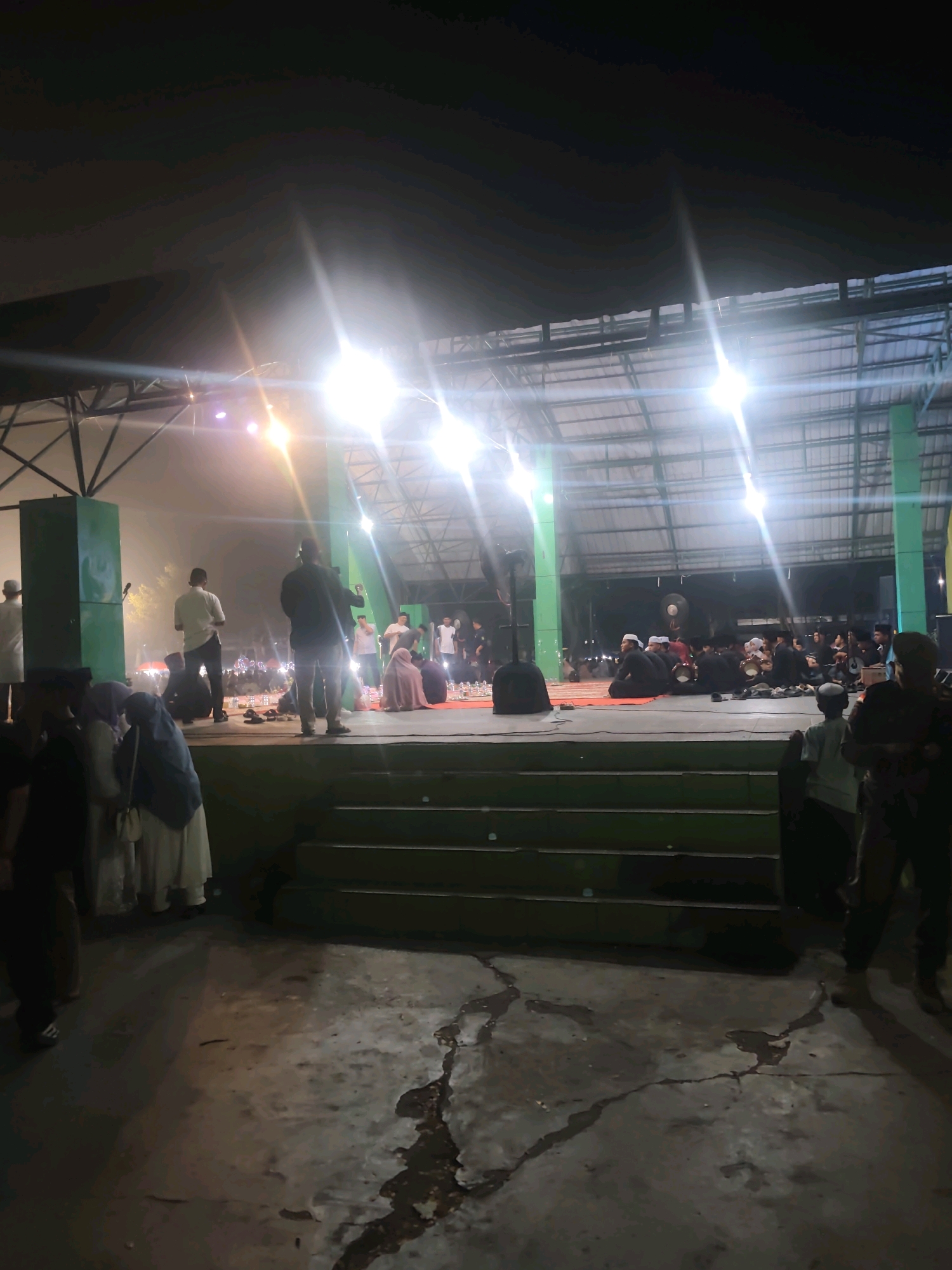 The image shows an outdoor stage at night in Rengat, Indonesia. Several people are visible on and around the stage. On the stage, there are individuals standing and sitting, some appearing to be performers or speakers. A crowd is gathered in front of the stage, some seated on the ground. The stage is illuminated by bright lights, creating lens flares. In the foreground, there are steps leading up to the stage, and the ground appears to be concrete with some puddles or stains. The overall atmosphere suggests a public event or performance taking place.