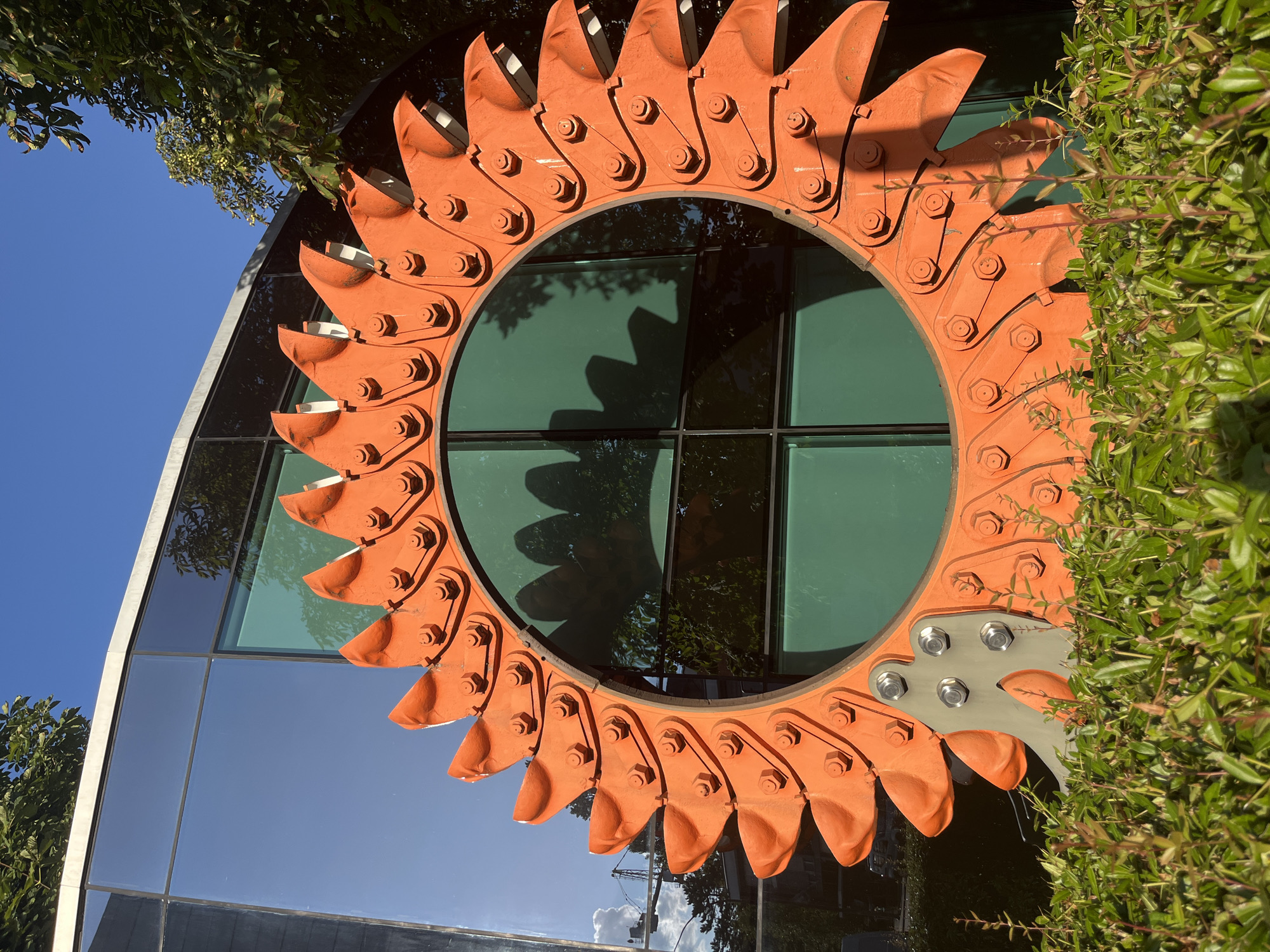 This is a medium shot, eye-level, outdoor photograph of a large, orange, circular metal sculpture with a saw-tooth edge. The sculpture is bolted to a silver metal base which is partially obscured by a dense, green hedge in the foreground. Behind the sculpture, the reflection in the glass windows of a modern building shows the sculpture itself, as well as trees and a blue sky with some clouds. The building's exterior is a combination of dark and green tinted glass panels. The sculpture is positioned in front of the building, with trees and the sky visible above and around it. The overall impression is that of a piece of industrial art situated in an urban or suburban environment. The sculpture is not identified as being in Geneva, Switzerland, as there is no information in the image to support this claim.