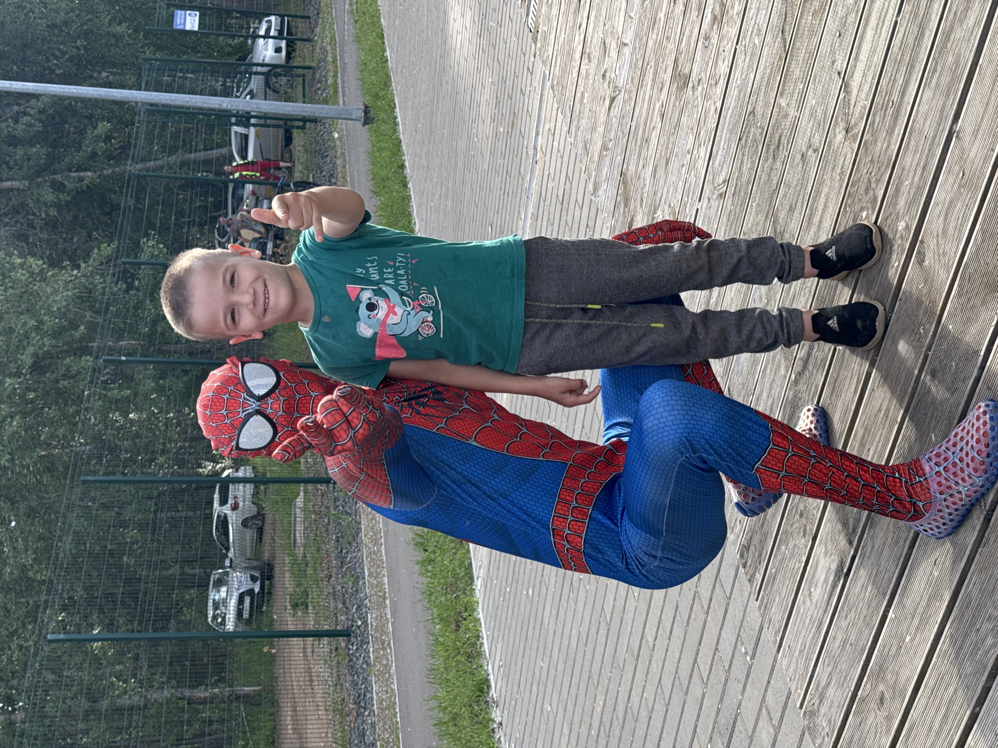 In a sunlit outdoor scene on a wooden deck, a young boy in a green t-shirt with a koala graphic gives a thumbs-up. He is positioned next to a person dressed in a full Spider-Man costume, who is leaning back on the deck. The Spider-Man costume is bright red and blue with the iconic web pattern. The boy wears grey patterned pants and black sneakers. In the background, a green metal fence separates the area from a grassy verge and a line of trees. Beyond the fence, several cars are parked, and a pathway is visible. The lighting suggests it is daytime. The overall atmosphere appears cheerful and playful, possibly at an outdoor event or gathering in Chaikovsky, Russia. The text on the boy's t-shirt includes "y unts ARE OALA-TY!".