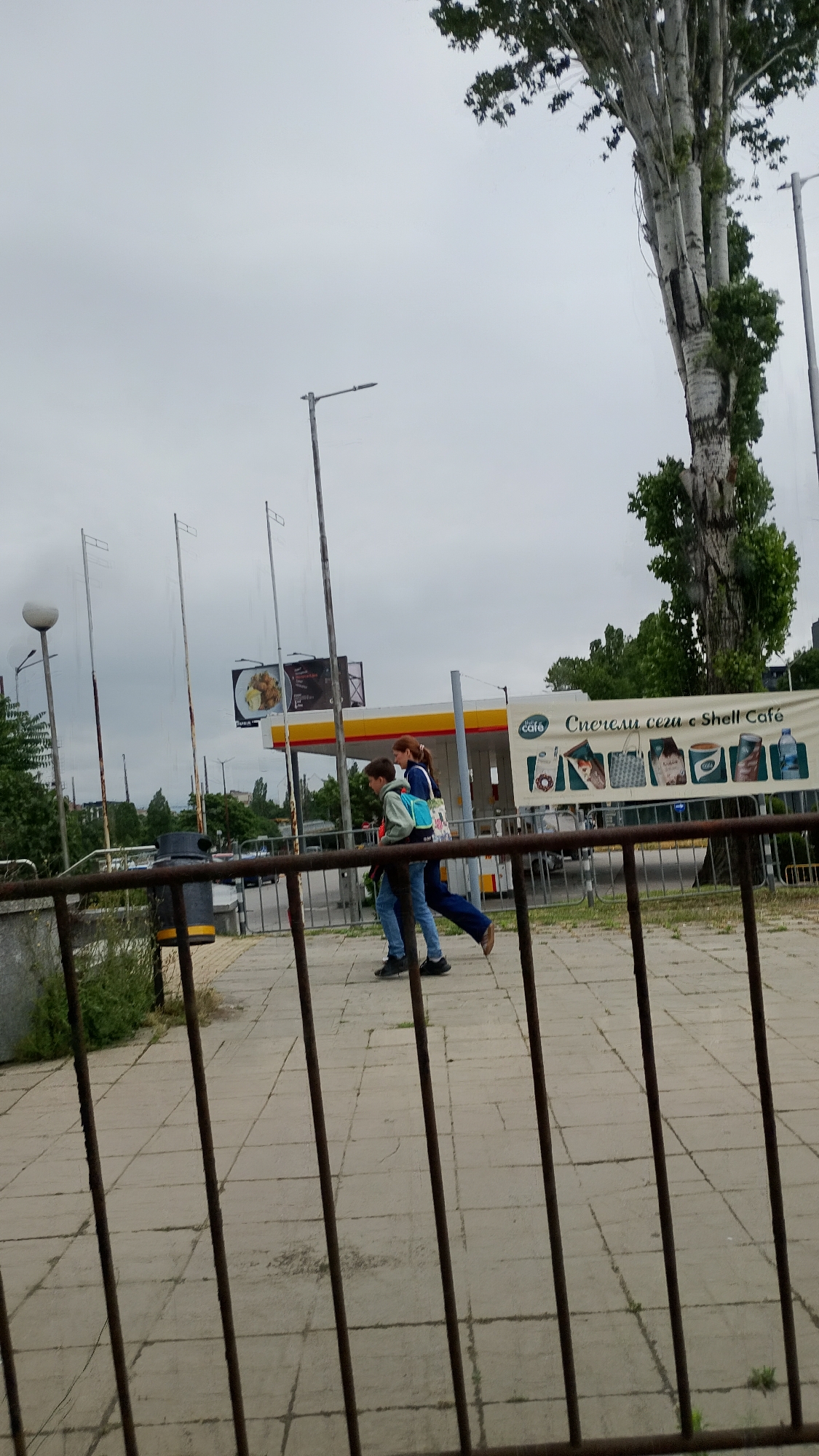 The image shows two individuals walking past a Shell gas station and cafe on a cloudy day. The foreground is dominated by a rusty metal fence, through which the pavement and a sidewalk are visible. Behind the fence, a gray trash bin with a yellow band is positioned near a low green bush. In the mid-ground, two people, one adult and one child, are walking from left to right. The child is wearing a light-colored hoodie and blue jeans, carrying a blue backpack. The adult is wearing a dark top and dark pants, also walking. The Shell gas station, with its distinctive yellow and red canopy, is visible in the background, along with a large billboard advertising food items and a banner for "Shell Café". Several tall streetlights are scattered across the scene. To the right, a large tree with a pale, mottled trunk stands prominently against the overcast sky. The environment appears to be an urban setting, likely a public area adjacent to a road or commercial establishment. Based on the "Shell Café" branding and the likely presence of Bulgarian language on the signage, the location is presumed to be Sofia, Bulgaria.