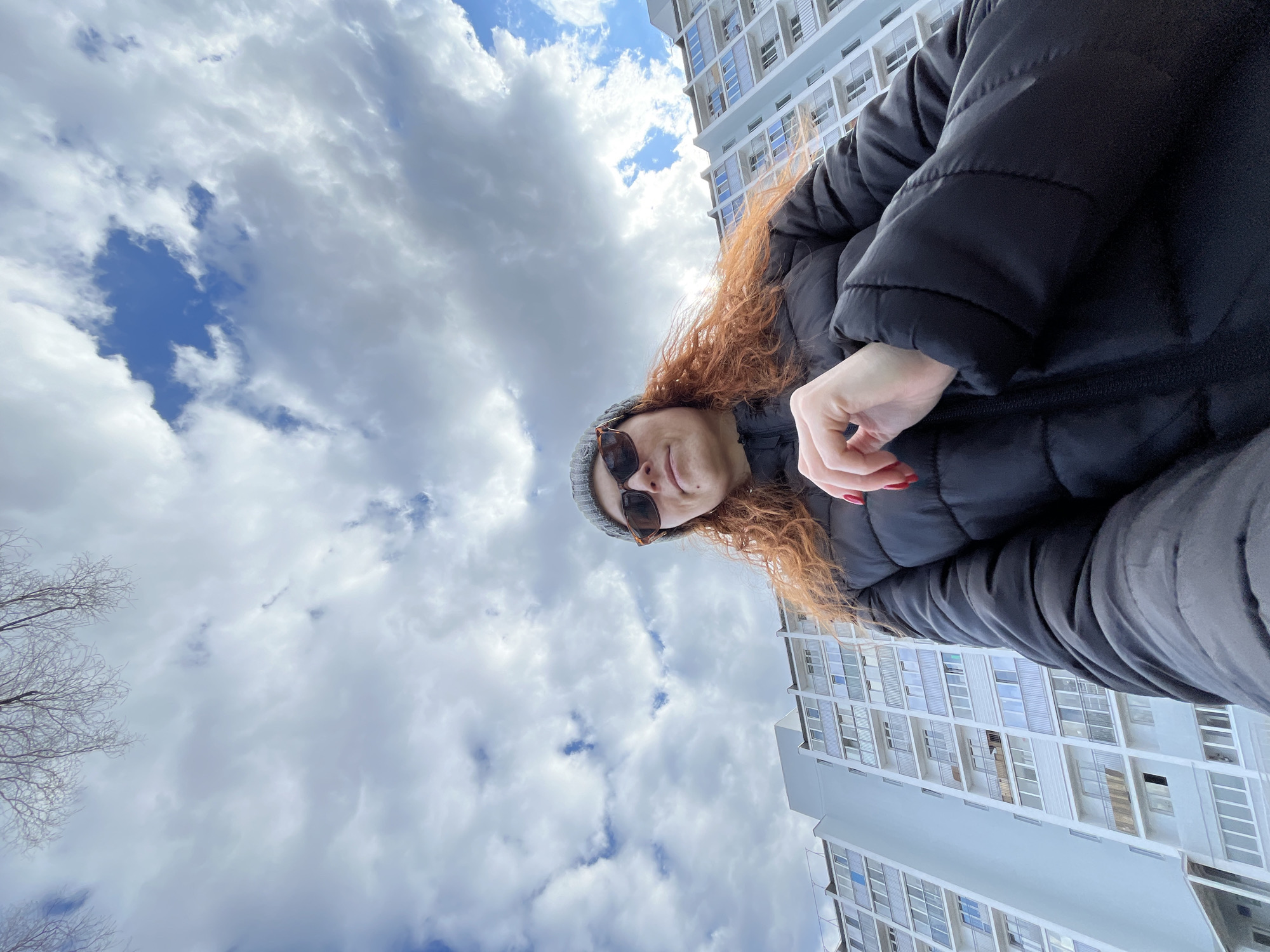 An outdoor, low-angle photograph captures a person taking a selfie, with a prominent sky and modern building as the backdrop in Chaikovsky, Russia. The main subject is an adult with long, curly, reddish-brown hair, wearing a dark (likely black) puffer jacket, a grey beanie hat, and sunglasses. Her lips are slightly parted, suggesting a relaxed expression or faint smile. Her left hand is visible, featuring red nail polish.
The camera is angled sharply upwards, showing the subject from below. The sky dominates the upper and left parts of the frame, filled with a mix of large white and grey cumulus clouds, interspersed with patches of clear blue sky. On the right side, a tall, light-colored, multi-story building with numerous windows and balconies rises towards the sky, indicating an urban or residential setting. Bare tree branches, characteristic of late autumn, winter, or early spring, are visible on the far left edge of the image.
The overall impression is of a bright, cloudy daytime scene. There is no visible text in the image. The unusual low-angle perspective provides an artistic and immersive feel to the outdoor portrait.