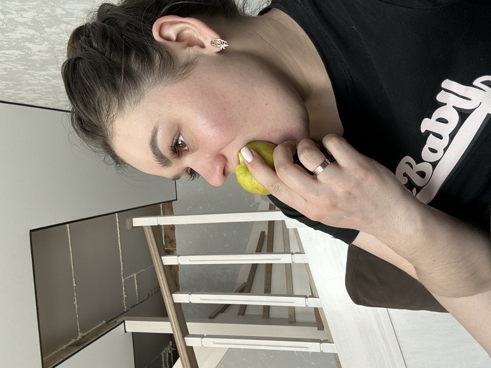 A woman is holding a green pear to her cheek. She is wearing a black t-shirt with the word "Baby" on it, and a gold ring on her finger. The setting appears to be indoors, possibly a home, with a partially constructed staircase visible in the background. The location is suggested to be Chaikovsky, Russia.