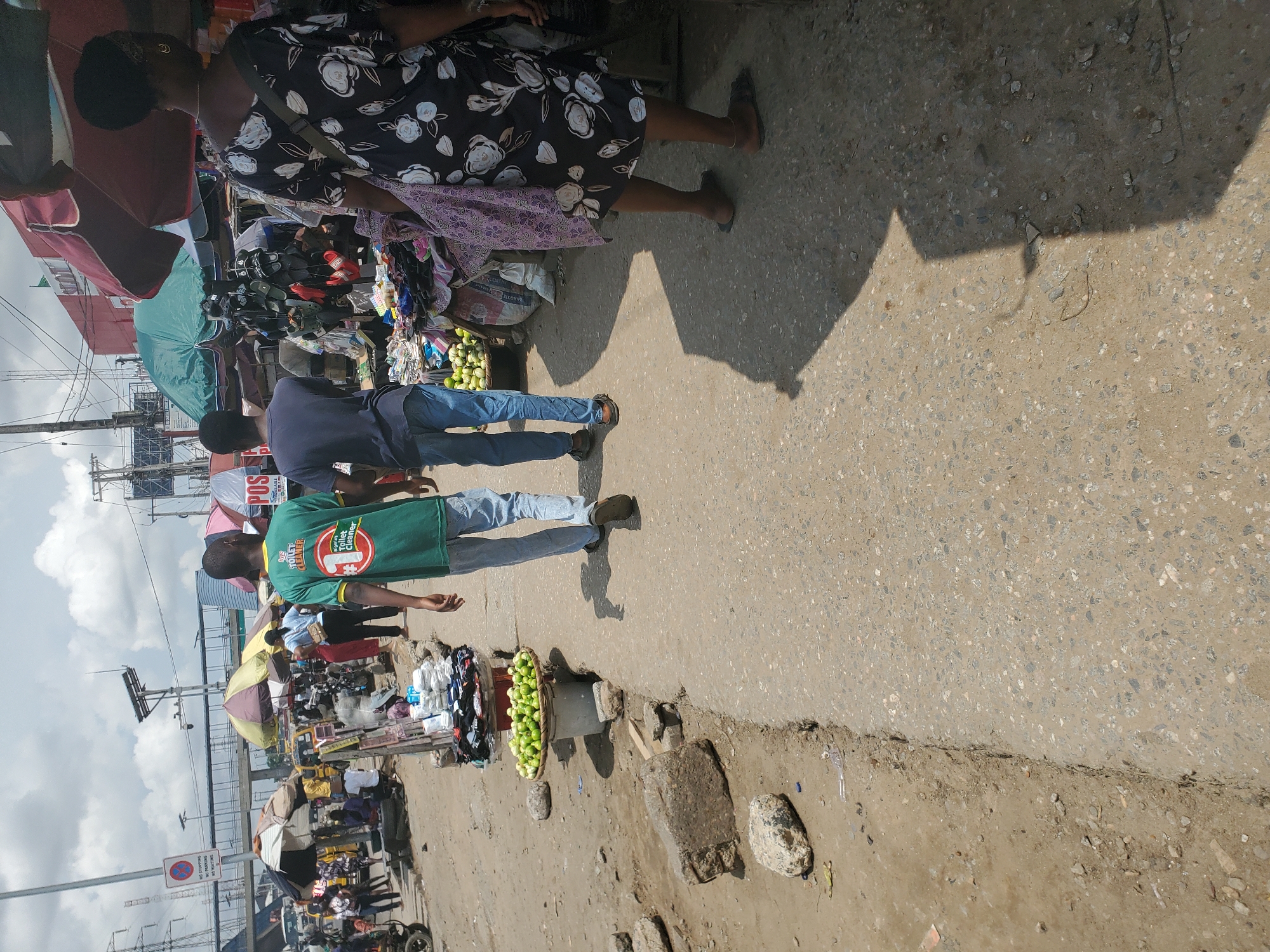 The image captures a bustling street scene in Shangisha, Nigeria, with an emphasis on commerce and daily life. Several people are visible, primarily focused on their activities within the market. In the foreground, two young men are walking, one wearing a green t-shirt with text that reads "Toilet Cleaner" and a graphic of a toilet. The other man is in a blue t-shirt. Behind them, a woman in a floral dress is seen in profile, appearing to be engaged in shopping or browsing.
The setting is an outdoor market with various stalls and displays. Numerous objects are scattered throughout the scene, including clothing, shoes, and a prominent basket overflowing with green fruits, likely limes or small melons. Umbrellas and awnings provide shade over some of the stalls, suggesting a sunny day. The ground is a textured concrete and dirt surface, typical of a vibrant market environment.
The overall atmosphere appears to be active and commercial, with people going about their business. The time of day is likely daytime, indicated by the strong shadows cast on the ground. There is a sign visible on a pole with text that reads "NO STOPPING" and "NO PARKING." The presence of stalls, goods for sale, and people actively participating in the market points to a typical day of commerce in this Nigerian location.