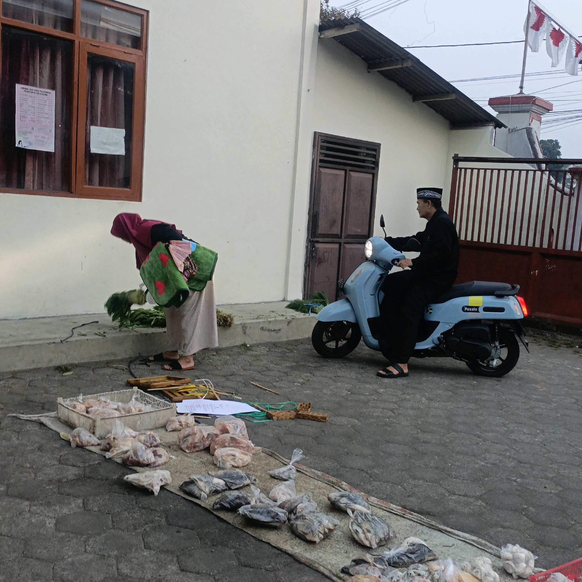 The image depicts an outdoor scene in Salatiga, Indonesia, likely a market or roadside stall, with a building in the background. A person wearing a dark Islamic cap and a black robe is riding a light blue scooter. In the foreground, a person wearing a maroon headscarf and a long skirt is bent over, appearing to tend to produce or other goods laid out on the ground. A variety of items, including what appear to be packaged meats and dried fish, are displayed on mats and in a crate. The building in the background has a window with brown trim and a corrugated metal roof extension. Flags are visible on a pole near the top right of the frame. The ground is paved with interlocking bricks.