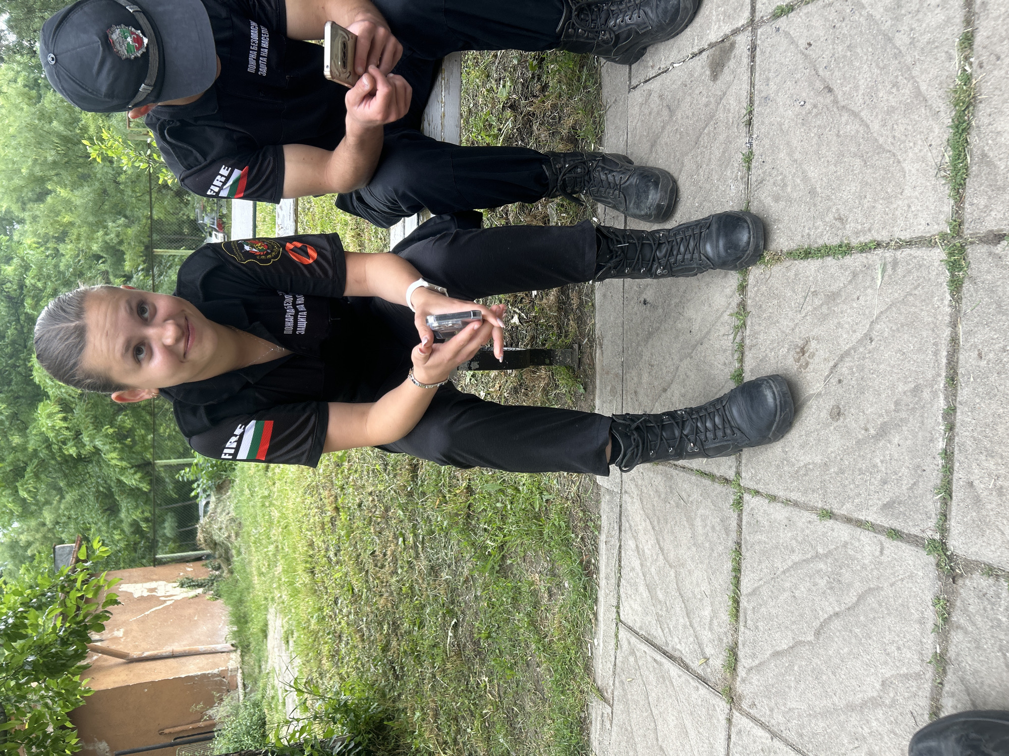 Two individuals in dark uniforms sit on a bench outdoors. The person in the foreground is a woman looking at the camera, holding a cell phone. Both are wearing black boots. The uniforms feature the word "FIRE" and Bulgarian flags. The location appears to be a park-like setting with greenery and tiled pavement. Given the Bulgarian flags on the uniforms, it is likely that the location is in Bulgaria. Assuming that this a common uniform for firefighters in Bulgaria, and given the prevalence of parks in the capital, I place this in Sofia, Bulgaria.