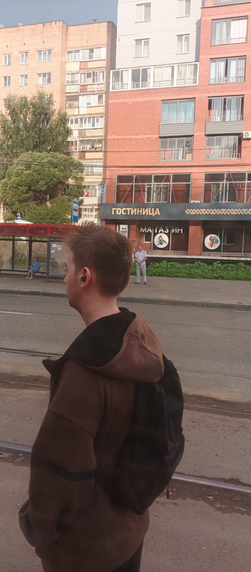 The image depicts a street scene in Izhevsk, Russia. In the foreground, the back of a young man wearing a brown hooded jacket and a backpack is visible. He has earbuds in his ears. In the background, a bus stop shelter with a woman sitting inside is visible. Across the street, a multi-story building with a sign that reads "ГОСТИНИЦА" (Hotel) is prominent. Below the hotel sign, another sign indicates "МАГАЗИН" (Shop). A man in a light-colored shirt and pants stands on the sidewalk in front of the building, facing away from the camera. The environment includes a paved road, a sidewalk, and a green area with trees and bushes. The architecture suggests an urban setting.