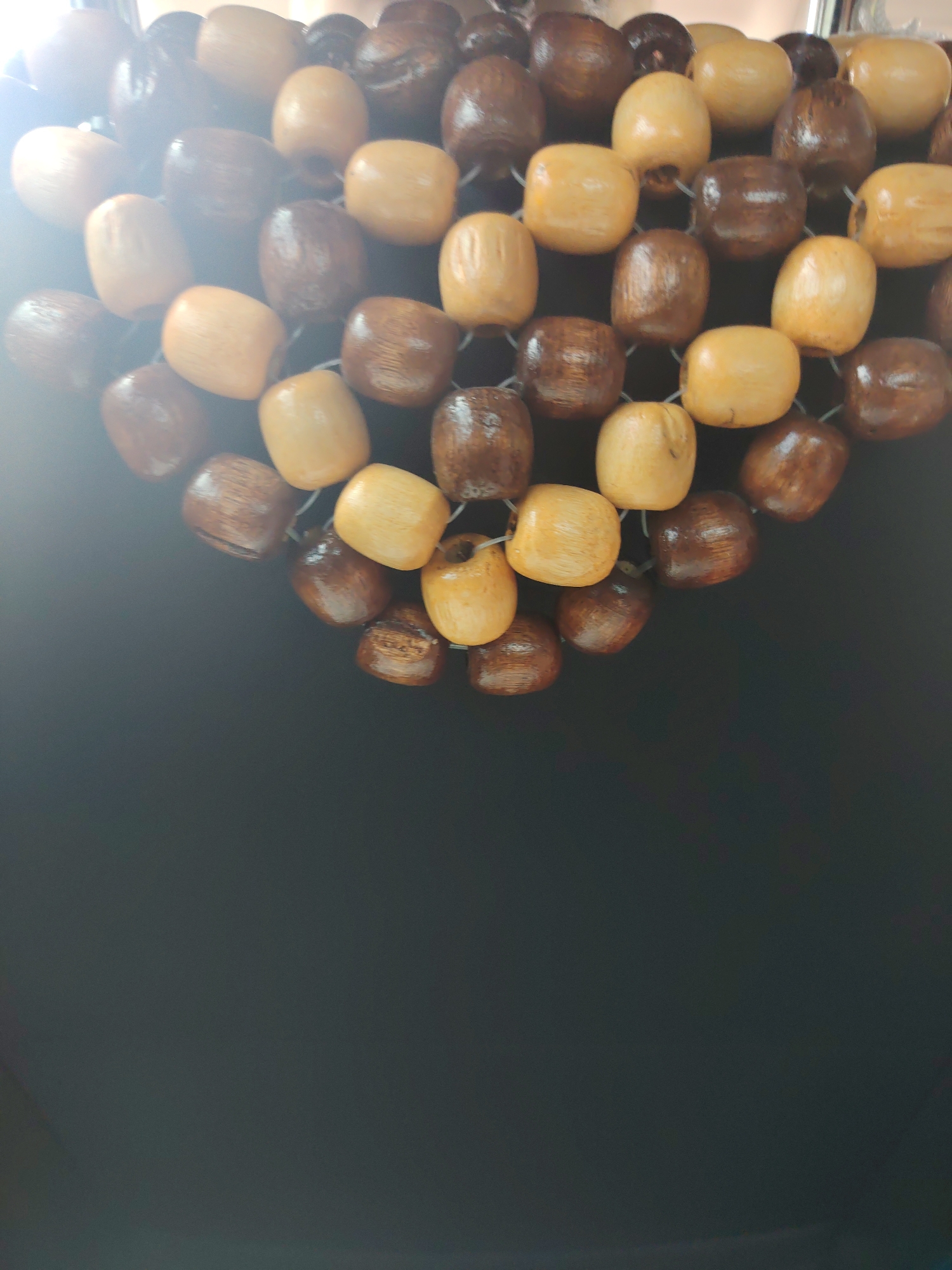 The image shows a close-up, overhead view of a beaded curtain or screen. The beads are made of wood and come in two colors: a light tan/yellow and a dark brown. They are oval-shaped with rounded edges and appear to be strung together on thin white cords, creating a mesh-like pattern. The beads are tightly packed and form a curved shape, suggesting they are part of a decorative item. The background is a plain, dark gray or black surface, out of focus. The lighting creates some glare and highlights on the polished surfaces of the beads. There are no people or any other discernible objects in the image.