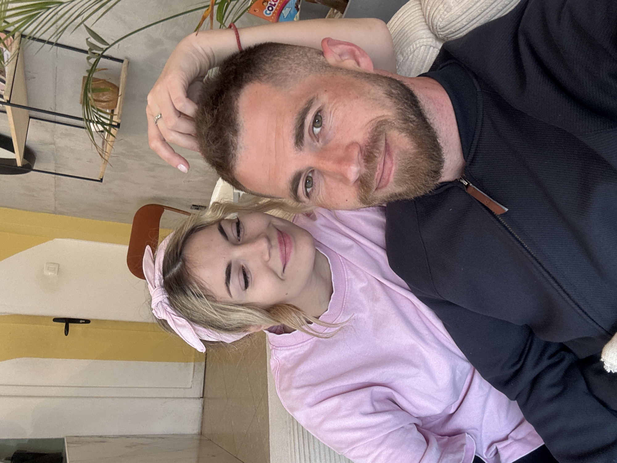 A couple is posing for a close-up selfie indoors. The man, on the right, has short brown hair and a beard and is wearing a black jacket. The woman, on the left, has blonde hair, a pink headband, and a pink sweater. The scene appears to be in an indoor setting, possibly a cafe or home, with a yellow and white wall in the background. There are plants in pots visible in the upper left of the frame. The specific location in Budva, Montenegro, cannot be determined from the image.