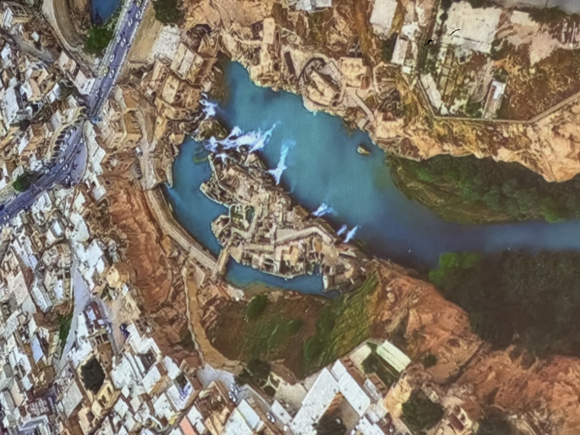 This is a high-angle, aerial-view photograph of an urban landscape with a river or body of water running through it. The water is a vibrant turquoise color and features several sections of white rapids, indicating a dam or waterfall.
The surrounding environment is a mix of natural and man-made structures. On the left side of the image, there is a densely populated urban area with many buildings. These buildings have light-colored walls and flat roofs, arranged in a somewhat irregular pattern. A road or pathway with parallel lines runs through this urban area, suggesting transportation infrastructure.
The center of the image is dominated by the water and several structures built within or around it. These appear to be ruins or ancient buildings, with a complex layout of walls and courtyards. The architecture suggests historical significance, possibly an old city or settlement.
To the right of the water, the landscape transitions to rugged, rocky terrain with patches of green vegetation, suggesting a more natural and uncultivated area. Further to the right, more buildings are visible, some with distinct white roofs.
The lighting suggests it is daytime, with shadows indicating the sun is not directly overhead. The overall scene is one of a historical settlement integrated with a natural water feature. There is no visible text in the image.