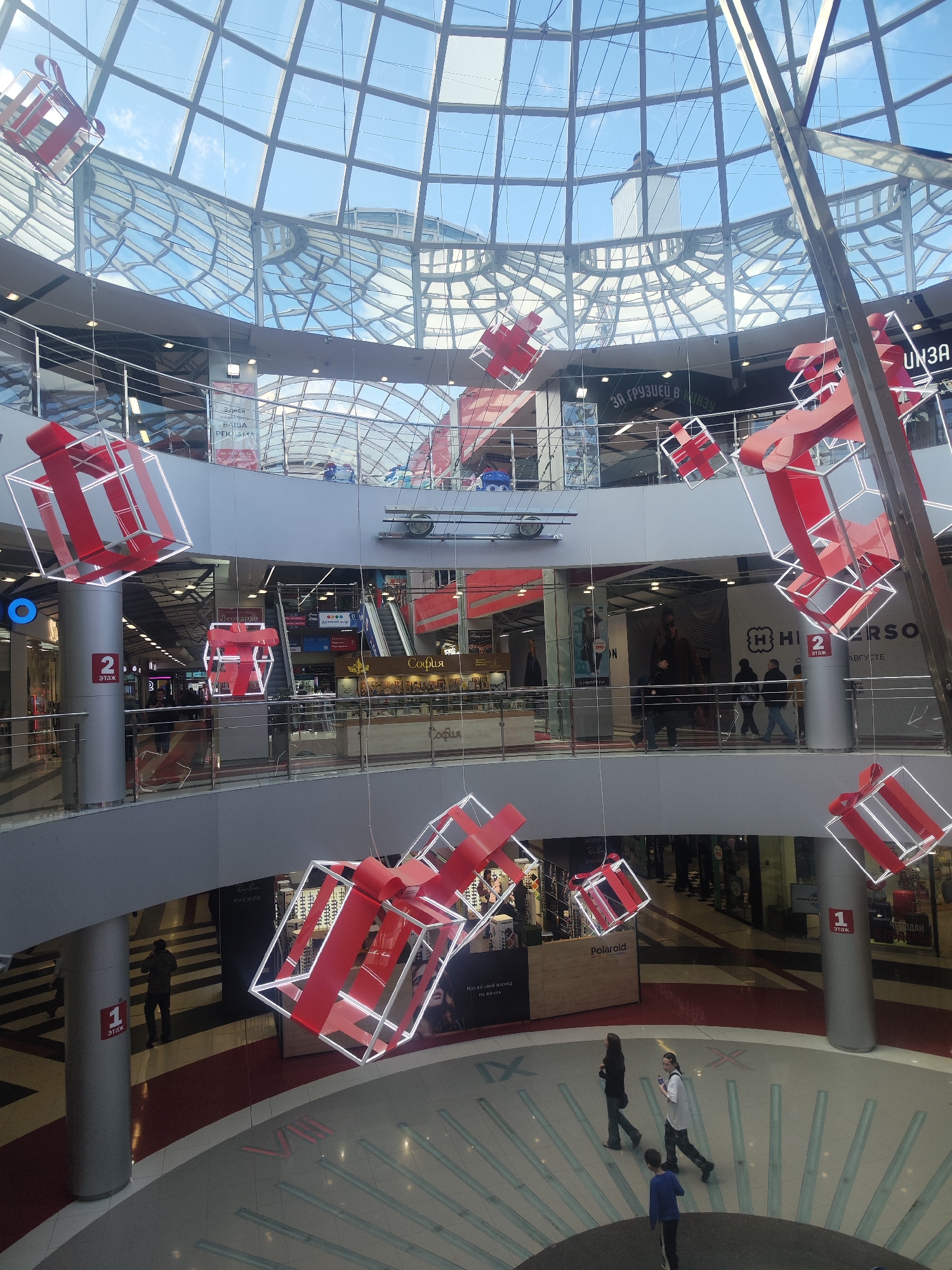 Captured from an elevated perspective, the interior of a bustling shopping center is illuminated by natural light filtering through a dome-shaped glass ceiling. A network of metal beams intersects across the dome, lending a structural element to the expansive overhead view. Multiple floors are visible, each lined with various retail stores and eateries, indicated by signage in Cyrillic script. Numerous individuals are seen strolling on the lower levels, and shoppers can be seen on the higher floors. Decorative, illuminated gift-box-shaped art installations, adorned with red ribbons, are suspended at different levels, adding a festive touch to the commercial space. Circular floor designs, reminiscent of a clock face, add a unique architectural detail to the interior. The location can be identified as the shopping center in Izhevsk, Russia.