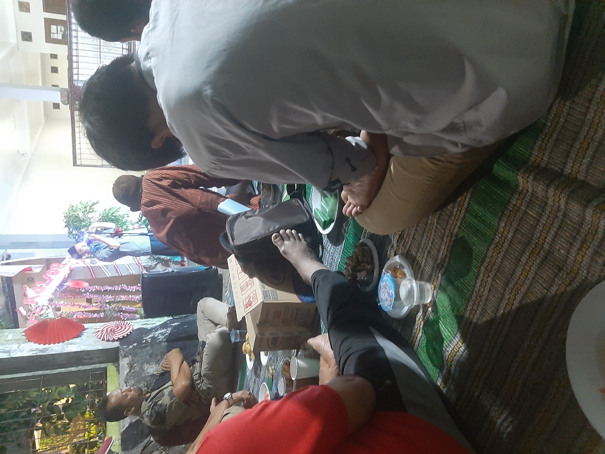 In the city of Sumampir, Indonesia, an outdoor gathering is taking place at night. Several individuals are seated on a woven mat on the ground, engaged in what appears to be a meal or a social event. In the foreground, a person wearing a red shirt is partially visible, facing away from the camera. To their right, another person is seated with their legs extended, wearing black pants and a grey shirt. Their feet are visible, with one resting on a beige-colored fabric.
Further back, a man in a patterned shirt and light-colored pants is seated with his arms crossed, looking to his left. Beside him, another individual is wearing a brown, corduroy-like shirt and a dark cap. In the background, a man in a light blue shirt and a dark cap stands near a decorated archway with red and pink ornaments and banners, seemingly addressing a group or participating in a ceremony. A black speaker is placed on the ground near the man in the brown shirt.
Various objects are scattered on the mat, including plates of food, plastic cups, and a cardboard box. The environment suggests a casual setting, possibly a backyard or community space, with a stone wall and greenery visible to the left. The lighting is dim, indicating nighttime, with some artificial lights illuminating the scene.