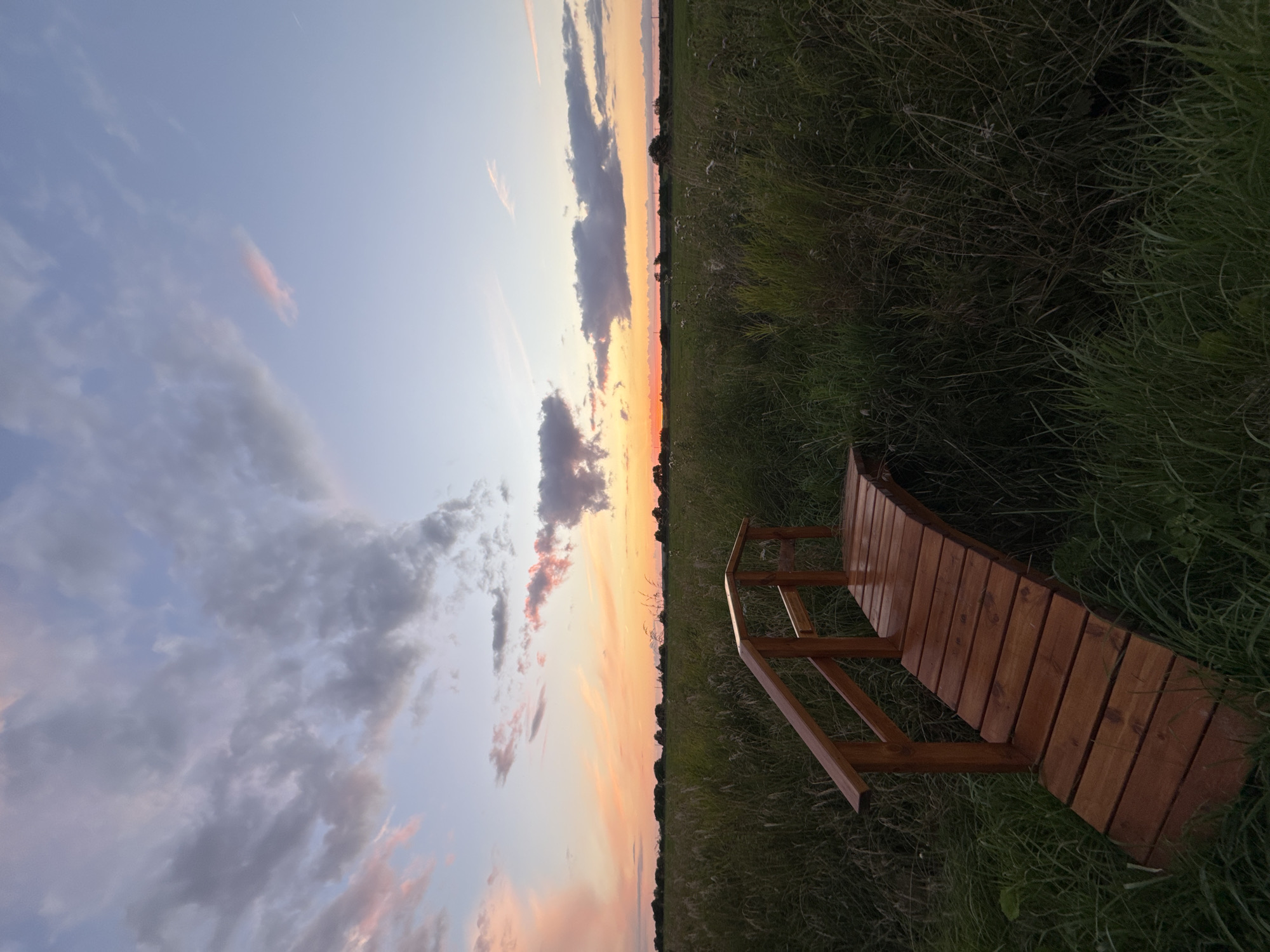 A wooden footbridge arches over tall grass in a field at sunset. The sky is a gradient of soft blues, pinks, and oranges, with scattered clouds. The field stretches to the horizon, dotted with trees. The bridge is constructed from polished wooden planks and features simple wooden railings. The lighting suggests early evening, with the sun low in the sky. The environment is natural and serene, characteristic of a rural or park setting. The image was captured in Kempelevo, Russia.
