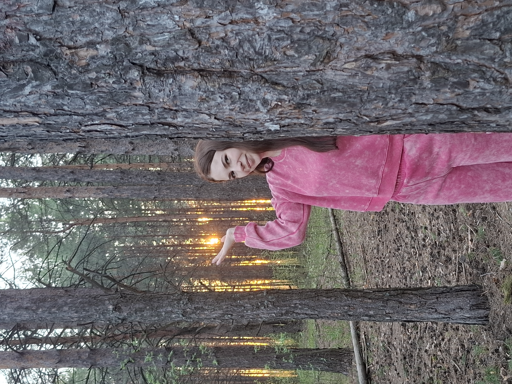 A young woman is standing among trees in a forest, likely in Chaikovsky, Russia, given the context. The woman has brown hair and is wearing a pink sweat suit. She is positioned between two large pine trees, appearing to interact with sunlight filtering through the trees. The environment is a typical forest setting, with numerous trees, ground foliage, and signs of sunset or sunrise. The overall impression is a natural setting with the subject interacting with the environment.