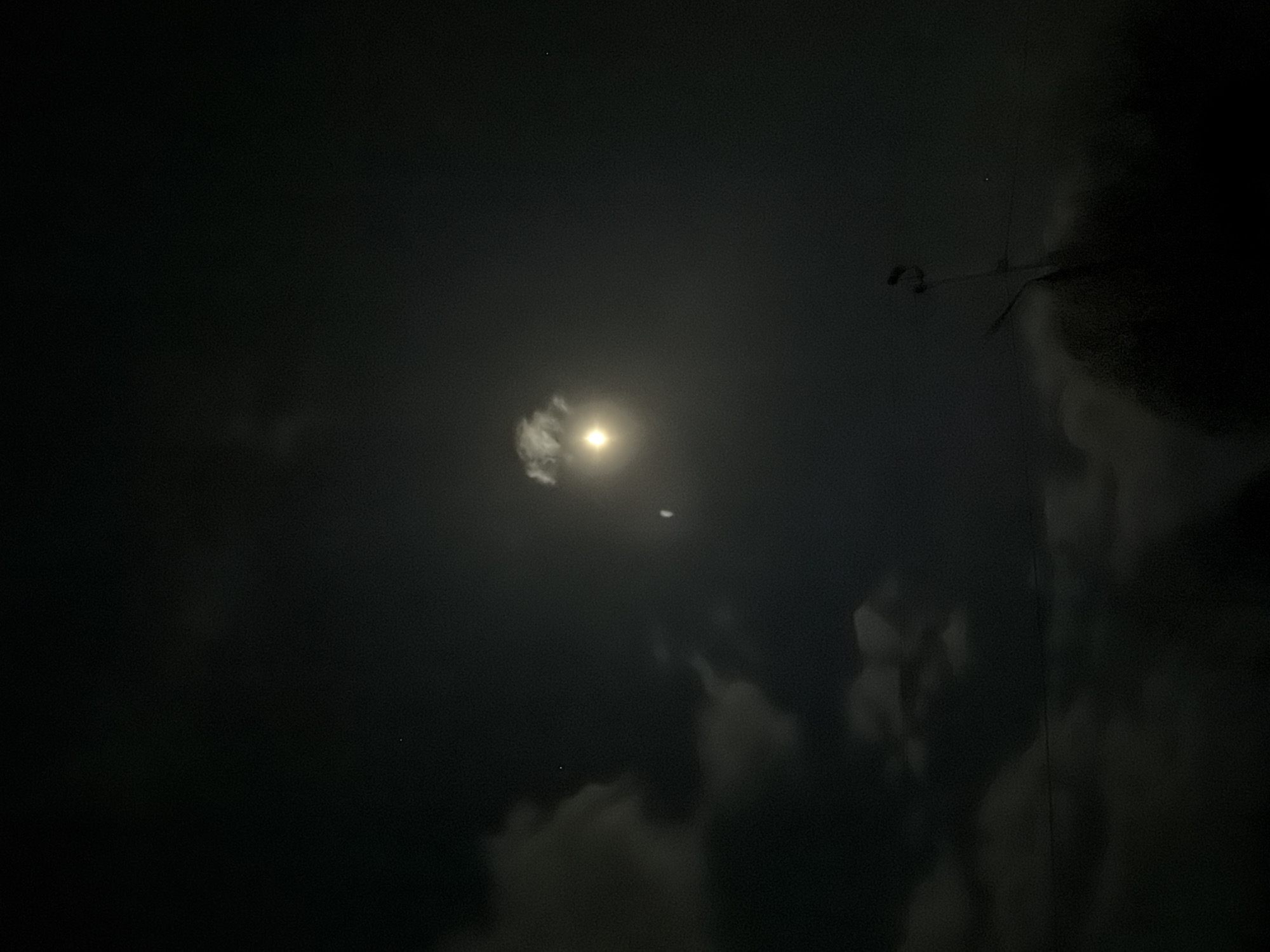 The image shows a night sky in Prikamskii, Russia. A bright moon is visible, surrounded by thin clouds. There is a small point of light below the moon, which might be a star or planet. Silhouetted against the sky, towards the right side of the image, is a blurred structure, potentially part of a building or utility pole. The overall scene is dark, with limited details visible beyond the moon and surrounding clouds.