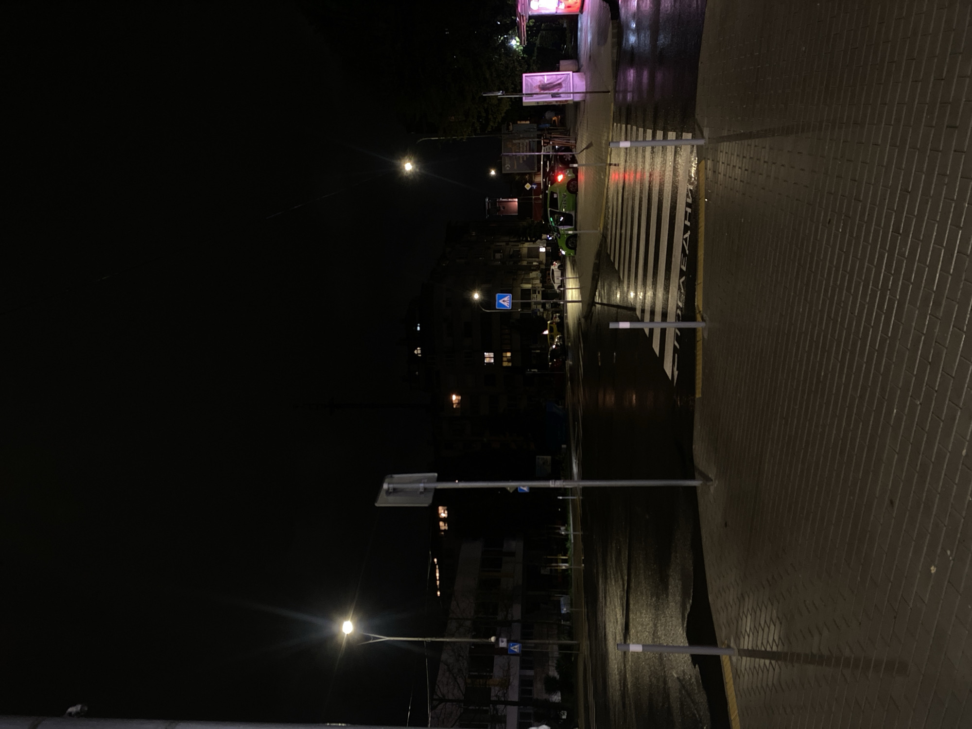 The image captures a nighttime urban street scene, likely after rain, given the wet and reflective surfaces of the road and sidewalk. The primary subjects are the street infrastructure and a few vehicles.
In the foreground and to the right, a tiled sidewalk made of dark grey pavers extends horizontally. To its left, a wide pedestrian crossing, marked with prominent white zebra stripes, stretches across the asphalt road. The road itself is dark and glossy, reflecting the artificial light sources.
Several streetlights are visible, casting pools of light onto the wet ground and illuminating parts of the scene. One bright streetlight is in the mid-ground on the left, and another is further back near the center. Various lampposts and traffic sign poles are distributed across the scene. A blue square sign with a white triangle depicting a walking person (a pedestrian crossing sign) is clearly visible near the center.
Further down the street, a few cars are parked or waiting. One car, predominantly green or yellow with white accents, is clearly distinguishable near the crosswalk, possibly a taxi or ride-share vehicle with faint text like "MAXI" on its side. In the distance, a red traffic light is visible. Behind the street and cars, apartment buildings with some illuminated windows are discernible against the dark night sky.
There are no people visible in the image. The time of day is night, emphasized by the darkness and reliance on artificial lighting. The wet conditions suggest recent rainfall.
Text is visible on the pedestrian crossing: "ПЕШЕХОДНА", written in white Cyrillic letters, which translates to "PEDESTRIAN". This detail, along with the urban architecture, is consistent with the provided location context of Burgas, Bulgaria. The overall atmosphere is quiet and still.