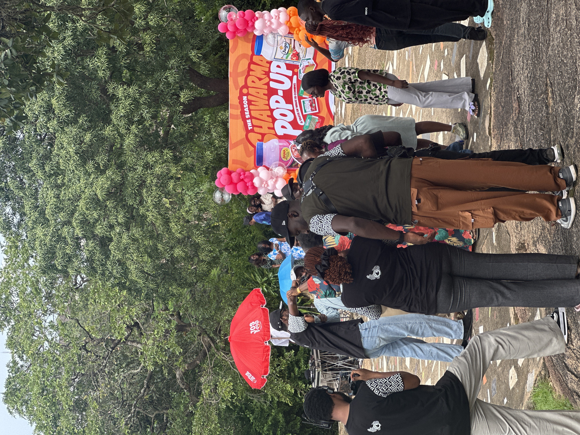 An outdoor event is taking place, likely a promotional pop-up, under a canopy of large trees. In the background, a large orange banner reads "THE SEASON SHARWARMA POP-UP A TASTE OF WHAT IS COMING." The banner is decorated with images of two jars of sauces, a tomato, and various food products. Pink and orange balloons frame the banner, and a few clear balloons are also visible.
A group of people are gathered in front of the banner. Most are casually dressed, with some wearing t-shirts and others in more formal attire. Several individuals are looking towards the banner or interacting with each other. A DJ setup with turntables and a mixer is visible to the left of the frame, under a large red umbrella with "JOS FOOD FESTIVAL" printed on it. A blue umbrella is also present.
The ground appears to be a mix of paved surfaces and natural rock formations. The overall atmosphere suggests a lively gathering, possibly a food festival or product launch, taking place in Jos, Nigeria. The presence of the "JOS FOOD FESTIVAL" umbrella strongly suggests the location is within Jos.