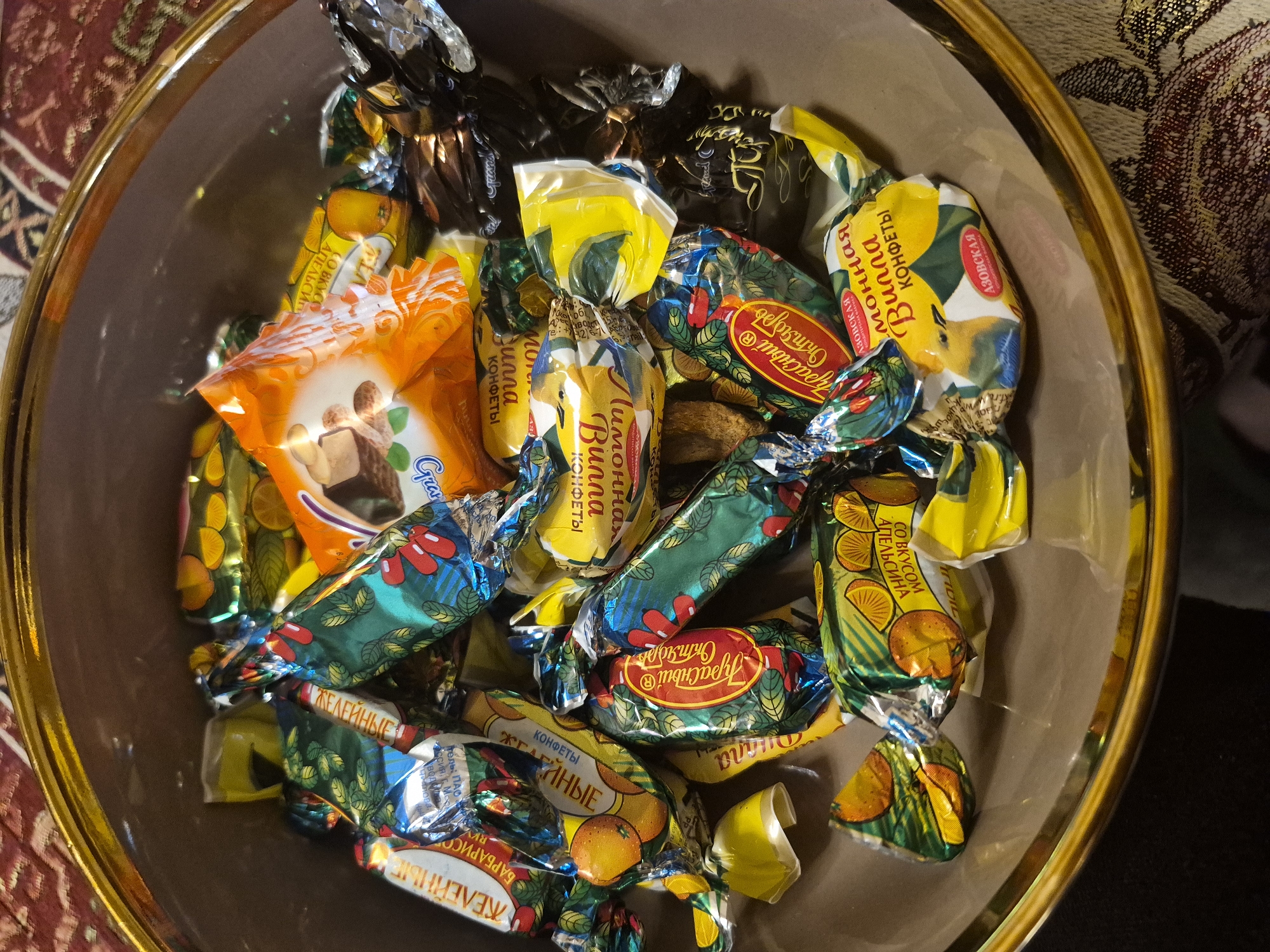A close-up, high-angle shot captures a brown bowl with a golden rim, brimming with a variety of individually wrapped candies. The candies are numerous and colorful, featuring wrappers in shades of yellow, orange, green, blue, and dark brown/black. Many wrappers are twisted at the ends, indicating individual packaging.
Visible text on the wrappers, primarily in Cyrillic, reveals several types and brands of confectionery. "АЗОВСКАЯ" (Azovskaya) appears multiple times, likely a brand name. Candies with yellow wrappers are labeled "Лимонная Вишня" (Lemon Cherry) and "КОНФЕТЫ" (Candies). Green wrappers with orange illustrations indicate "СО ВКУСОМ АПЕЛЬСИНА" (With Orange Flavor). Other wrappers are marked "ЖЕЛЕЙНЫЕ" (Jelly) and "БАРБАРИСО" (Barberry), suggesting jelly candies and a barberry flavor. A prominent red oval label on some green wrappers reads "Красный Октябрь" (Krasny Oktyabr), a known confectionery brand. An orange wrapper shows the word "Grand".
The candies fill the bowl, creating a vibrant, textured mound. The bowl itself is placed on a patterned fabric surface, possibly a tablecloth or upholstery, suggesting an indoor setting. The overall impression is one of a sweet treat offering. The image was taken in Yerevan, Armenia.