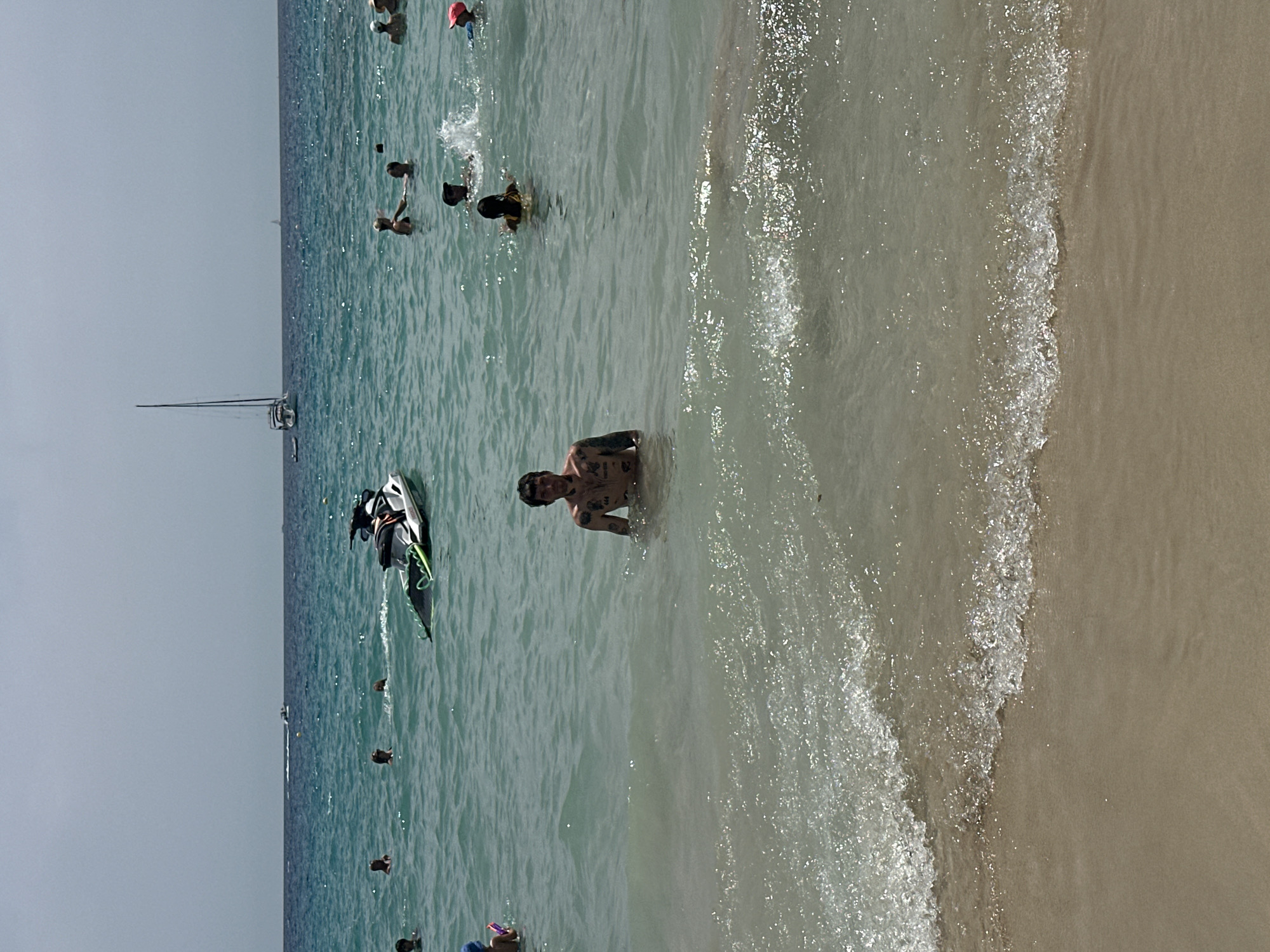 The image depicts a crowded beach scene with numerous people swimming and enjoying the water. In the foreground, a man with numerous tattoos is partially submerged in the ocean, looking towards the camera. Several other individuals are visible in the water, some interacting with each other. A jet ski is parked near a boat in the mid-ground, and a sailboat with a tall mast is visible in the distance. The environment is a sunny beach with clear blue water and a sandy shoreline. The location is the beach in Sant Agustí, Spain.