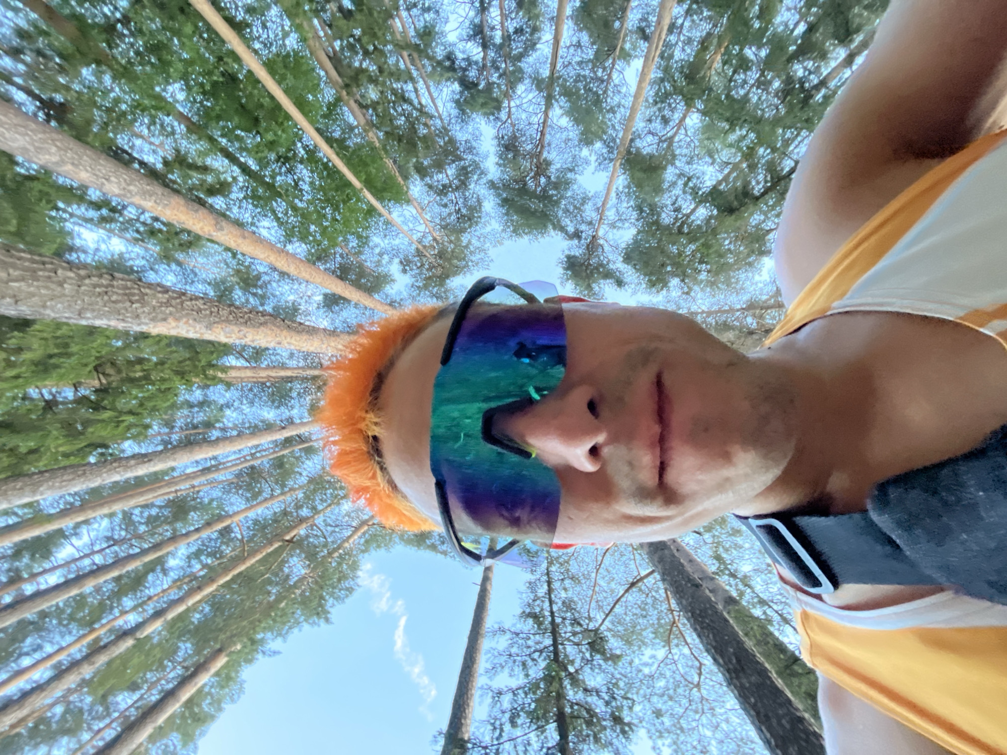 A low-angle, fisheye perspective selfie shows a person with bright orange hair wearing reflective, colorful sports sunglasses. The person is also wearing a yellow and white tank top and a black strap across their chest. The background is dominated by tall, slender pine trees reaching towards a bright blue sky with scattered white clouds. The image appears to be taken outdoors, possibly in a forest or park environment. No specific location in Chaikovsky, Russia is discernible from the visual information.