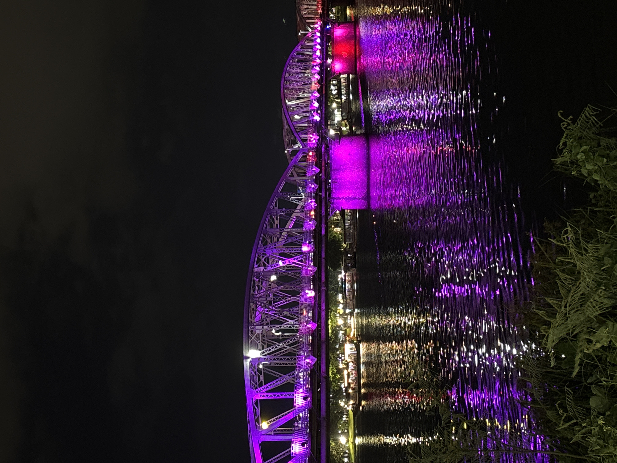 The image depicts a bridge illuminated with purple lights at night, spanning across a body of water. The bridge appears to be a truss bridge, with intricate metalwork visible. The water reflects the vibrant purple lights, creating a dazzling effect. In the background, there are faint lights suggesting a cityscape or buildings. Along the riverbank, there are trees and some vegetation in the foreground. Several boats with lights are also visible on the water, suggesting they are moored or navigating near the bridge. The overall scene conveys a sense of nighttime urban beauty and tranquility. The bridge is identified as the Trường Tiền Bridge in Phú Xuân Ward, Vietnam.