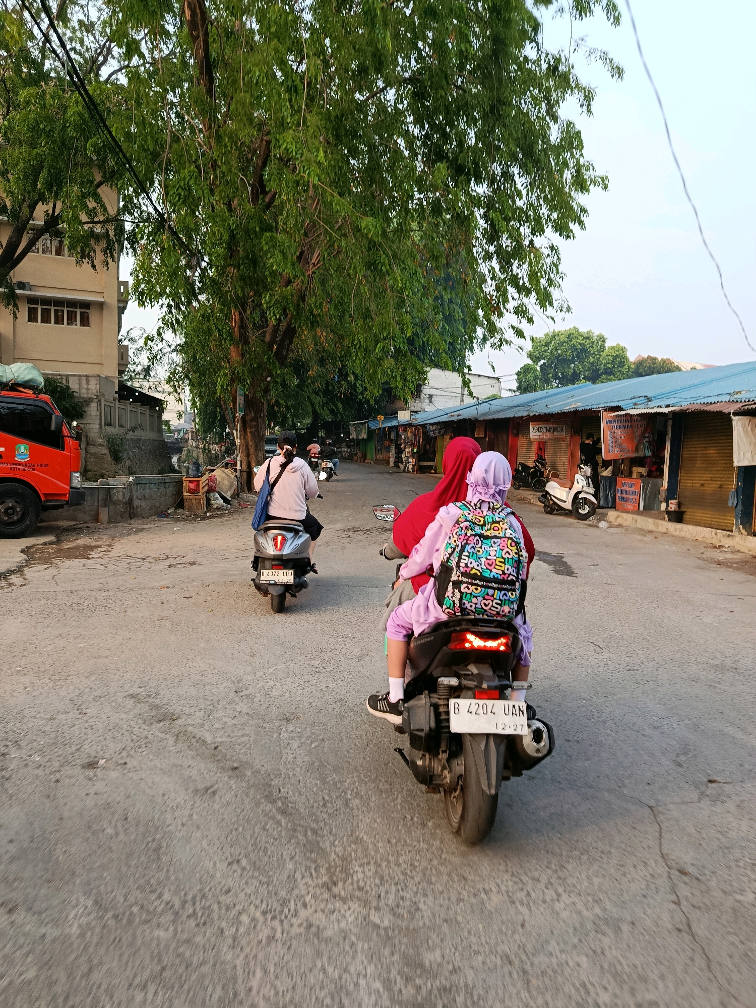 The image depicts a street scene in Bekasi, Indonesia, characterized by its urban environment and common modes of transportation. In the foreground, two individuals are riding on a motorcycle, viewed from behind. The rider is wearing a red headscarf and a light pink hoodie, while the passenger, a child, is wearing a purple hijab and a backpack adorned with colorful letters and patterns. They are traveling down a paved road lined with small shops and stalls, many of which appear to be closed or have their shutters down.
On the left side of the road, an orange truck is parked. Further down the street, other motorcycles can be seen, with one rider in a light pink hoodie and black shorts on a silver scooter. The buildings in the background are a mix of commercial structures with blue awnings and a taller, light-colored building with windows. Lush green trees with dense foliage are prominent throughout the scene, suggesting a warm climate. The sky is clear and bright, indicating daytime. The road surface is asphalt, with some visible cracks and markings. The overall impression is of a bustling, everyday street in an Indonesian city.