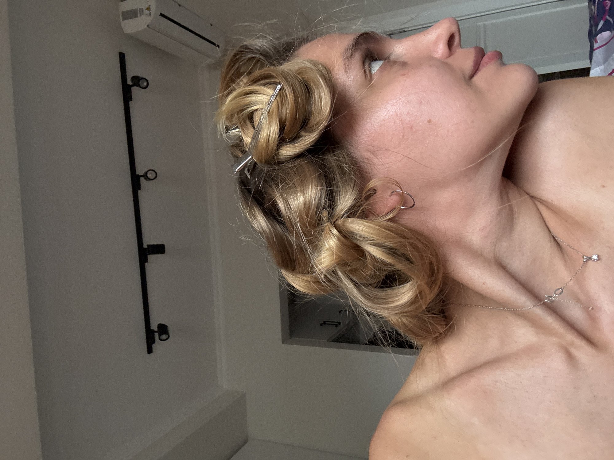 An indoor, eye-level shot features a person, likely female, from the chest up, with her head tilted sharply upwards and to the right. Her blonde hair is styled in sections, rolled into pin curls and held in place by several silver hair clips. She wears small, silver hoop earrings and a delicate silver necklace with two small charms, one appearing to be a bow with a tiny diamond-like embellishment. Her skin tone is light, and faint freckles are visible on her cheek and neck. Her eyes are looking upwards.
The background consists of a light-colored wall. In the upper left corner, a white air conditioning unit is mounted, with some text possibly reading "INVERTER" partially visible but not fully legible. Below it, a vertical black bar with two small black spotlights is attached to the wall. The lower right background shows a partial glimpse of what appears to be another white wall or door frame. The lighting suggests an indoor setting, and the activity appears to be related to hair styling. No strong emotions are discernible from the person's neutral expression.