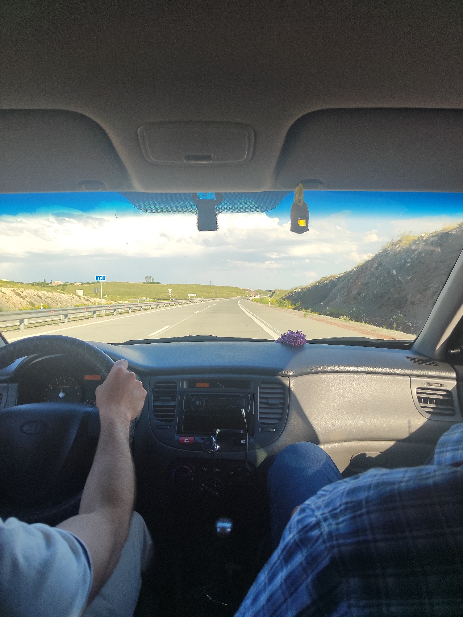 The image shows the interior of a car on a highway, likely during the daytime. The view is from the perspective of a passenger looking forward, with the driver's hands visible on the steering wheel. The dashboard, including the instrument cluster, air vents, and a car radio, is in focus. Outside the car, a highway stretches into the distance, with rolling hills and vegetation on either side. The sky is blue with scattered clouds. A speed limit sign indicating "110" is visible on the side of the road. There is a car air freshener hanging from the rearview mirror. A small bunch of purple flowers is placed on the dashboard. The overall impression is of a road trip or journey.