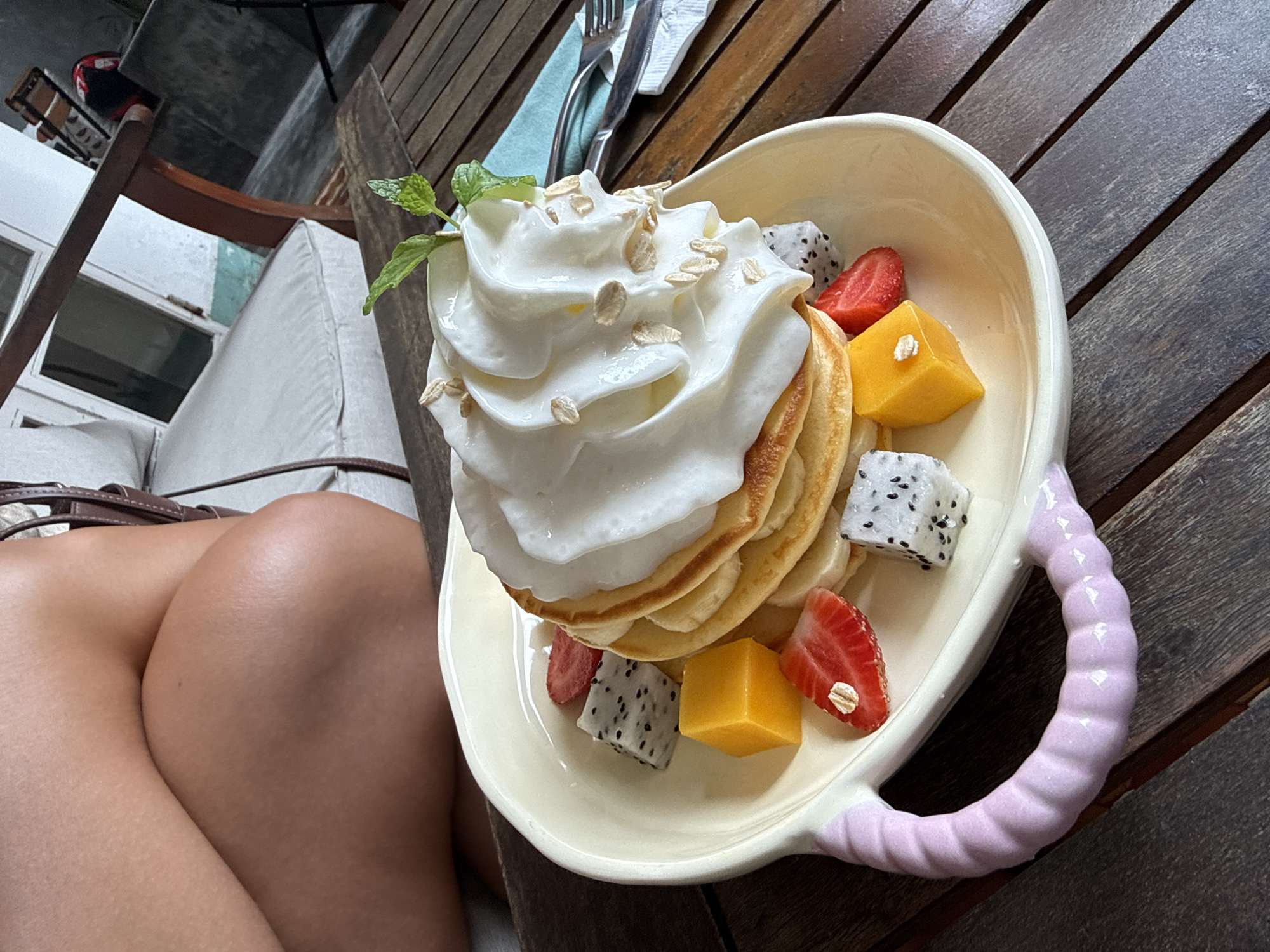 The image shows a plate of pancakes topped with whipped cream, oats, and various fruits (strawberry, mango, dragon fruit) on a wooden table. The plate is a light color with a textured pink handle. A person's leg and part of their buttock is visible in the lower left corner. Cutlery is present in the upper part of the image. A partial view of a cushioned seating area is in the background. The setting is outdoors, likely a cafe or restaurant. There is no confirmed information this image was taken in Da Nang, Vietnam, as there are no landmarks or distinct markers to pinpoint a precise location.