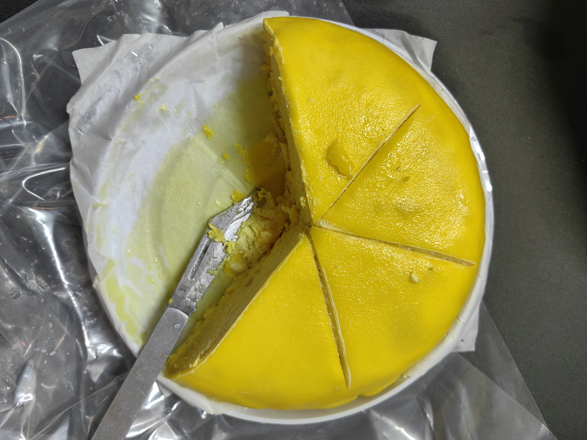 The image shows a cut yellow cake on a white plate, with a portion removed using a metal knife. The cake is divided into several slices. The plate sits on a surface that appears to be partially covered by a clear plastic bag or wrap. The background is a dark gray or black color. The location is not discernible from the image and the location provided cannot be confirmed from the given image.