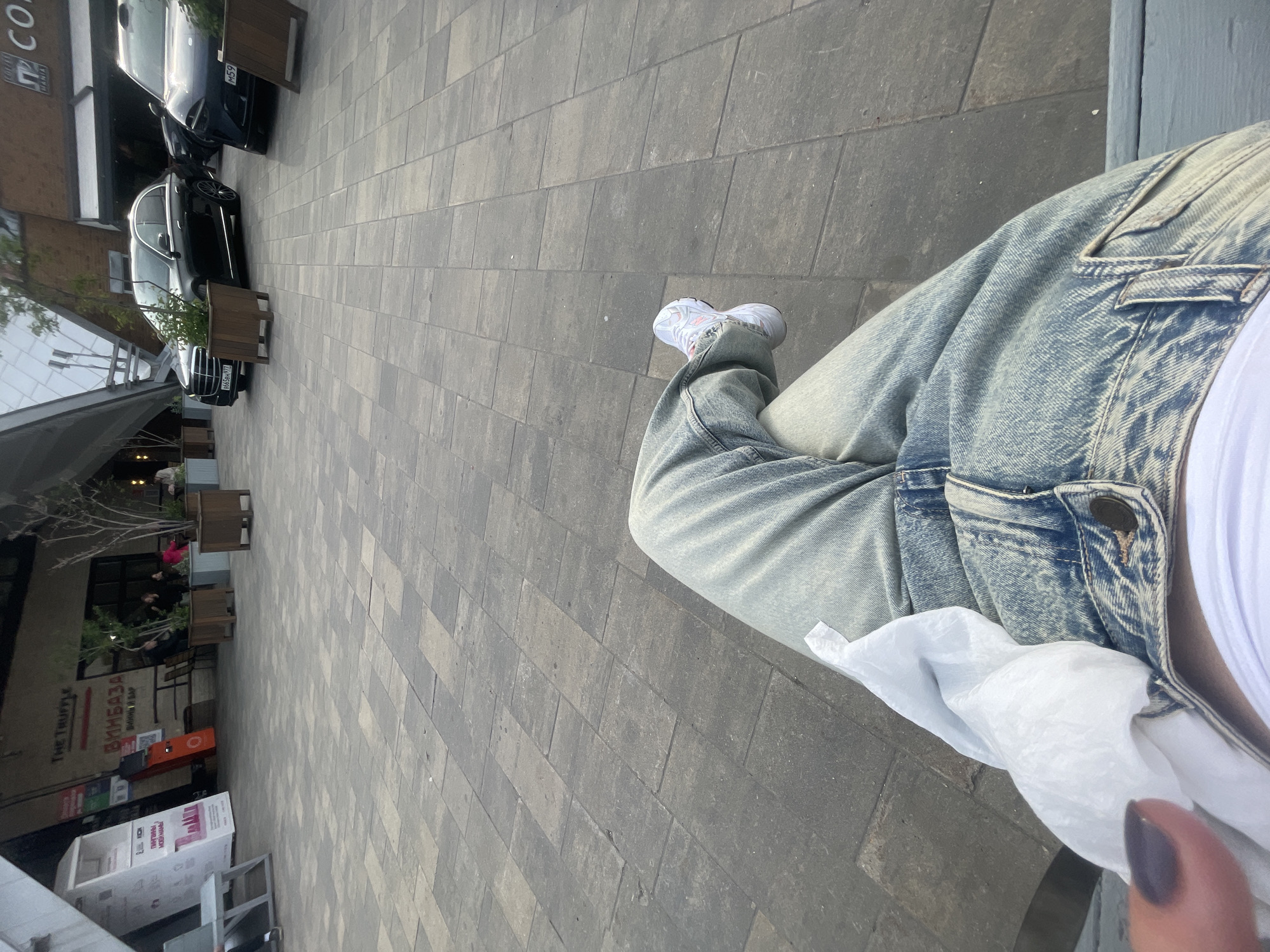 The image, taken at an oblique angle from a first-person perspective, shows a person sitting outdoors in Moscow, Russia. The individual is wearing acid wash jeans, a white shirt, and white sneakers. A portion of their finger with a dark nail polish is visible. They appear to be holding a white tissue.
The background shows a paved area, part of an outdoor commercial space. On the left side of the frame, there's a building with a sign that reads "THE TRUFFLE" and "ВИНБАЗА - ВИННЫЙ ВАР". There are also informational stands, potted trees, and cars parked nearby. License plates visible on the vehicles read "B665BB 977" and "M59". The overall environment suggests an upscale or modern commercial district.