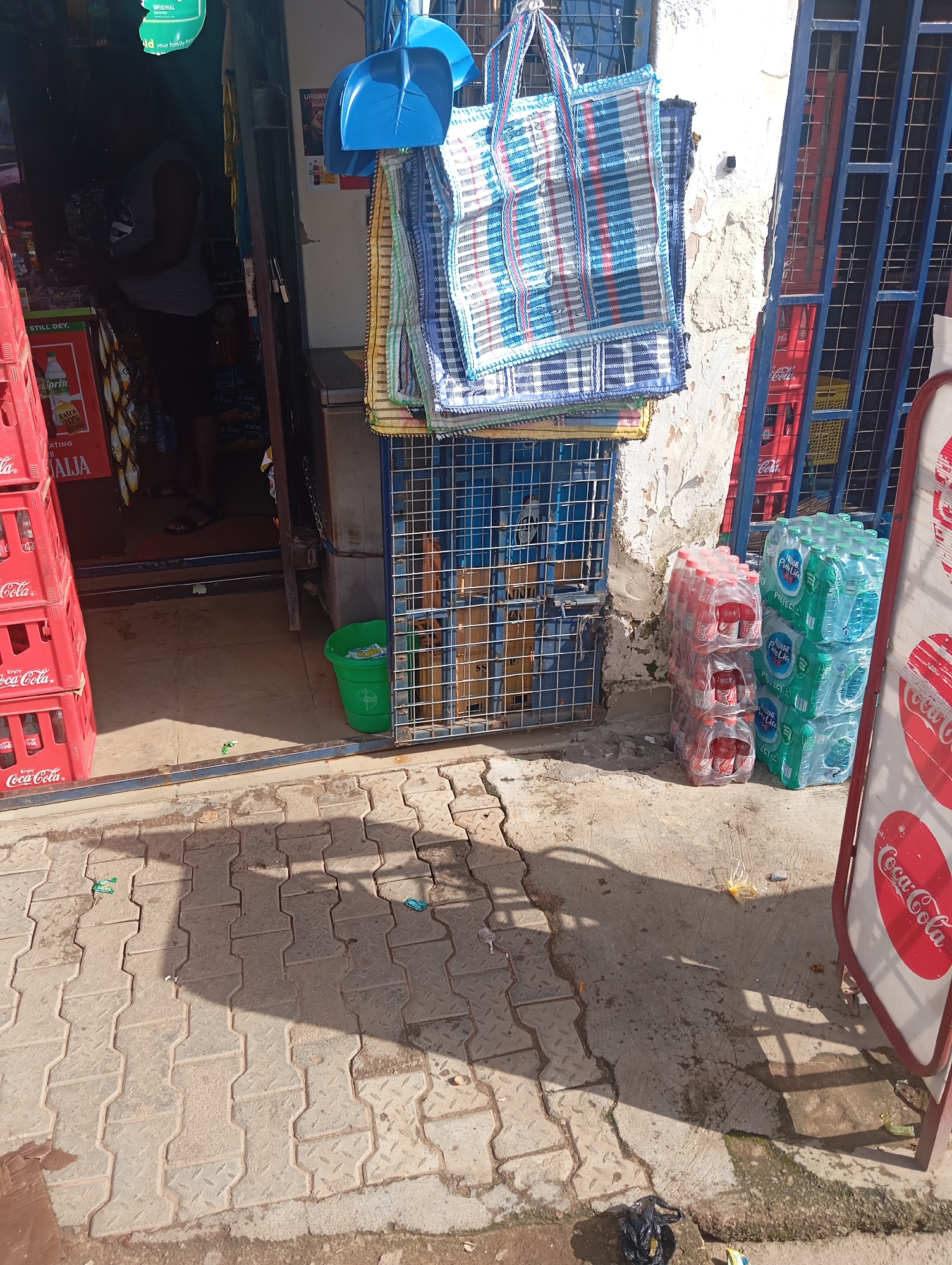 The image depicts the exterior of a small shop, likely a convenience store or kiosk, in Gura Top, Nigeria. The shop is open to the street, with goods displayed both inside and outside. In the foreground, stacked red Coca-Cola crates are visible, along with packs of bottled water labeled "Pure Life." A blue metal gate or shelving unit is positioned to the right of the entrance.
Hanging from the top of the entrance are two blue dustpans. Below these, several woven tote bags in blue, white, and red plaid patterns are displayed. Inside the shop, a person is partially visible, silhouetted against the interior light, appearing to be an employee or customer. Shelves with various products, including what appear to be snacks and drinks, are discernible within the dimly lit interior.
The ground outside the shop is paved with interlocking concrete blocks. The lighting suggests it is daytime, likely mid-morning or afternoon, with strong shadows cast on the ground, indicating a sunny day. There is some litter scattered on the pavement, including small plastic wrappers and a black plastic bag. A green bucket is placed near the entrance.
Visible text includes "STILL DEY," "ORIGINAL," "Id your family f," "UPDAT DAT," "Extra 20%," "ATING AIJA," "Coca-Cola," and "Pure Life." The overall scene conveys a typical street-level retail environment in Nigeria.