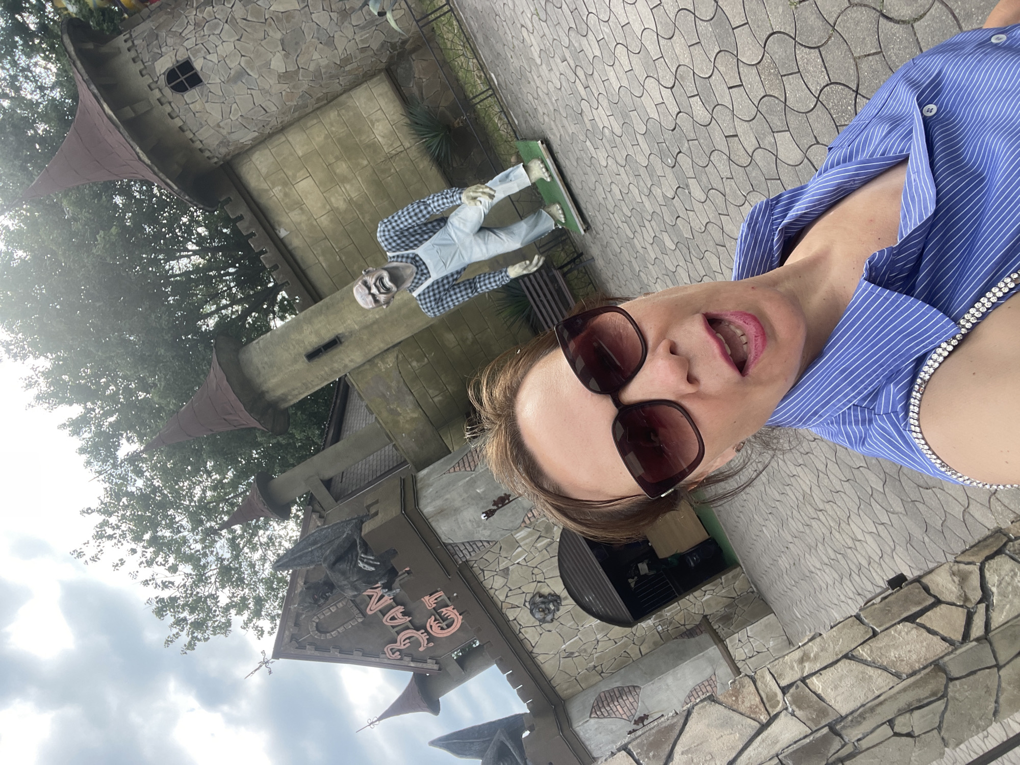 This is a selfie taken outdoors in front of a building with a medieval castle theme. The building is constructed from stone and features turrets with conical roofs. A sign above an entrance reads "Bagan", though the letters are stylized and somewhat obscured. A mannequin dressed as a zombie stands on a platform near the building.
The selfie is taken from a high angle, looking down at the woman. She is wearing a blue and white striped shirt and sunglasses. Her mouth is open, as if speaking or laughing. The ground is paved with interlocking stones. The weather appears to be overcast, with a grey sky visible in the background. The greenery of trees is also present.
The location is identified as Sochi, Russia. The overall atmosphere suggests a theme park or tourist attraction, likely related to fantasy or the macabre, given the zombie mannequin. The time of day is not explicitly discernible, but the lighting suggests daytime.