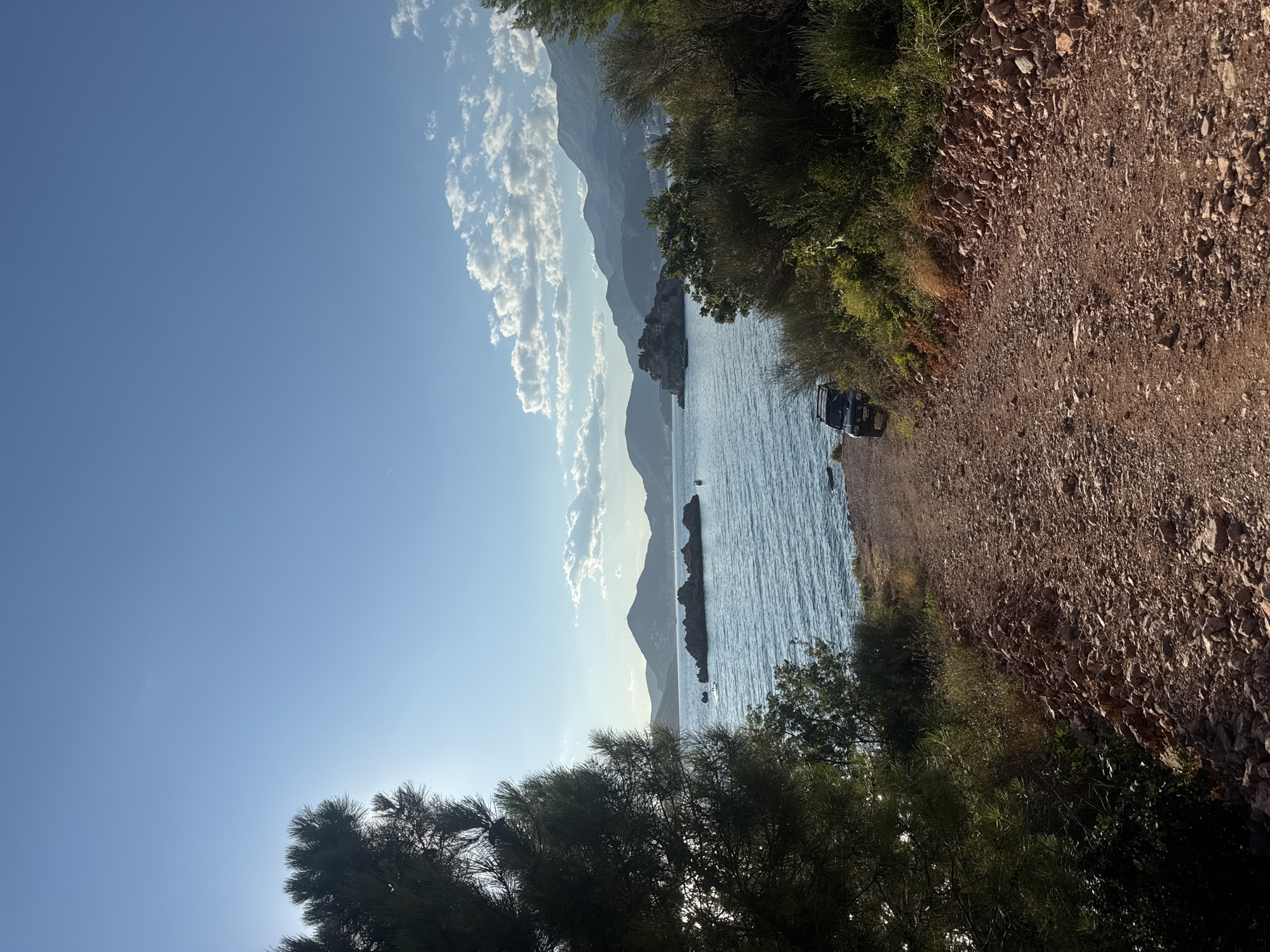 The image captures a scenic view of the Adriatic Sea from a dirt road in Sveti Stefan, Montenegro. The road, covered with reddish-brown gravel and stones, leads downhill towards the water. On the right side of the road, lush green vegetation and bushes grow, with some dry grass interspersed. On the left, a large pine tree with dark green needles is prominently featured, partially obscuring the sun which is low in the sky, casting a bright glare.
In the mid-ground, the calm blue sea stretches out. Several rocky islets, topped with vegetation, dot the water. In the distance, a chain of mountains forms the horizon. A small island, resembling the islet of Sveti Stefan, is visible on the left side of the frame, with buildings clustered on it and connected to the mainland by a causeway. A few small boats are present in the water.
A dark-colored, off-road vehicle is parked on the side of the dirt road near the bottom left of the frame, facing away from the viewer. The sky is a clear, bright blue with a few scattered white clouds in the distance. The overall impression is one of a tranquil, natural landscape bathed in the warm light of late afternoon or early morning.