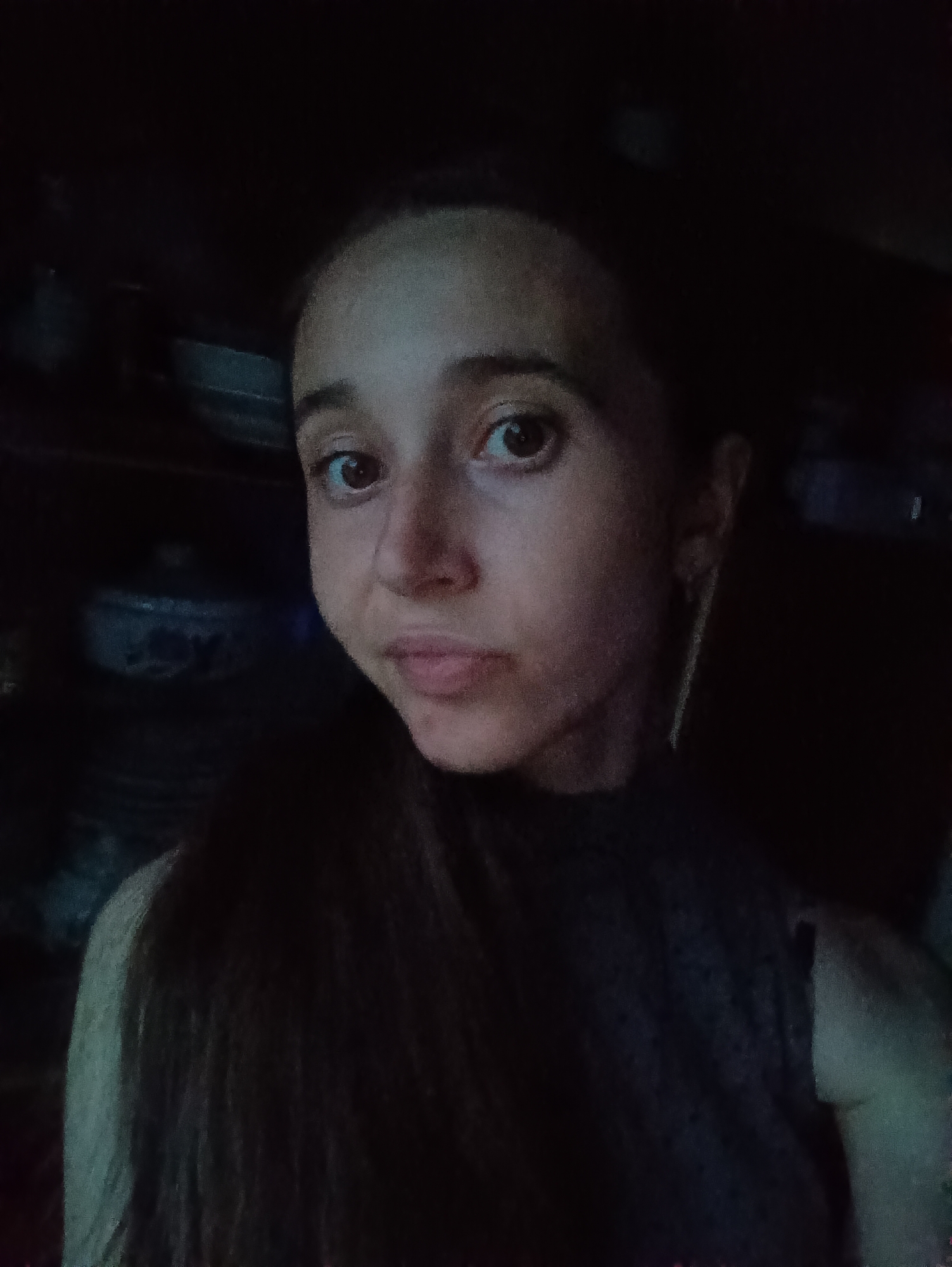 In Vypasne, Ukraine, a close-up selfie shows a young woman with long, dark hair and brown eyes. She is wearing a dark, sleeveless top. The background is dark and indistinct, with shelves of what appear to be bowls or pots visible. The lighting is dim, casting shadows on her face and the surroundings.