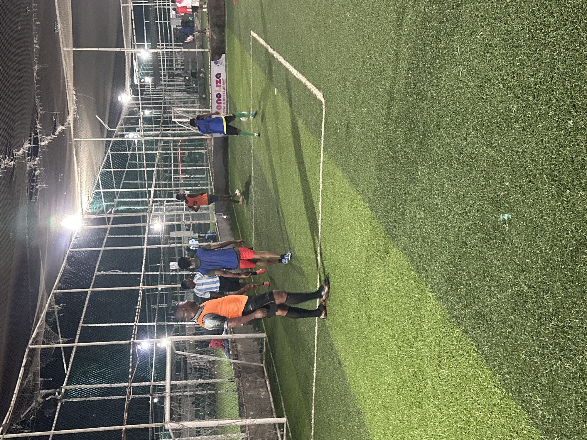 The image shows an artificial turf football pitch at night, illuminated by overhead lights. The pitch is fenced with netting and metal poles, typical of indoor or covered football arenas. Several individuals are present on the pitch, engaged in a game. One man in the foreground wears an orange bib over a grey shirt and black shorts, with black socks and orange and black football boots. To his left, another man wears a blue sleeveless top over a white and blue striped jersey, with red shorts and blue football boots. Further back, a man in a blue jersey and yellow shorts with green socks is kicking a football. Another individual in a red jersey with white trim and white shorts is visible on the right side of the pitch. In the background, more people can be seen sitting or standing outside the playing area. A sign that reads "Ono Liza Gym & Restaurant" is visible on the right side of the image, suggesting the location of this facility. The general atmosphere is that of a recreational football game taking place in an urban setting.