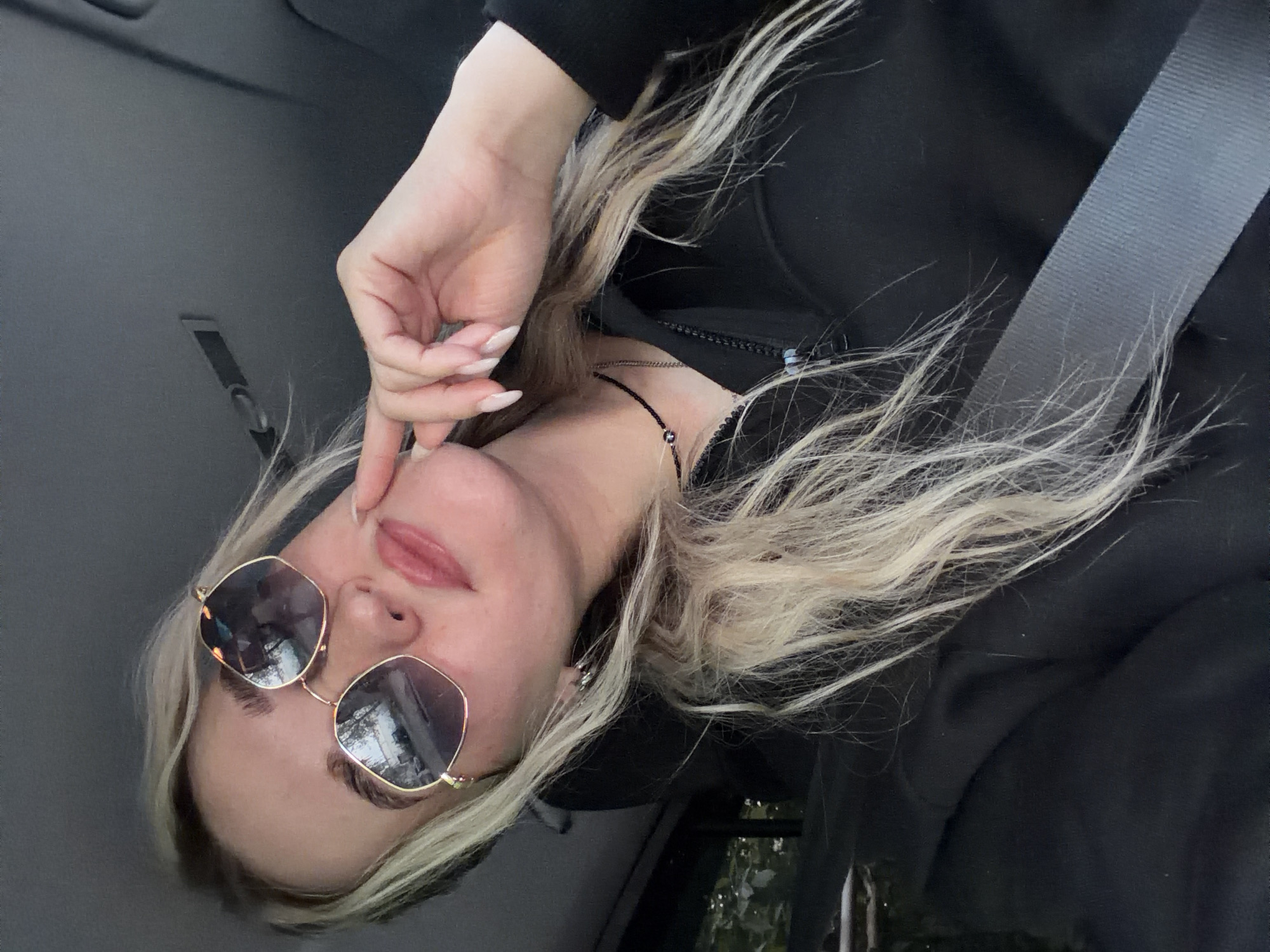 A person with blond hair and sunglasses is seated in a car. They are wearing a black sweatshirt and a seatbelt is visible. The location is Unknown, Unknown.