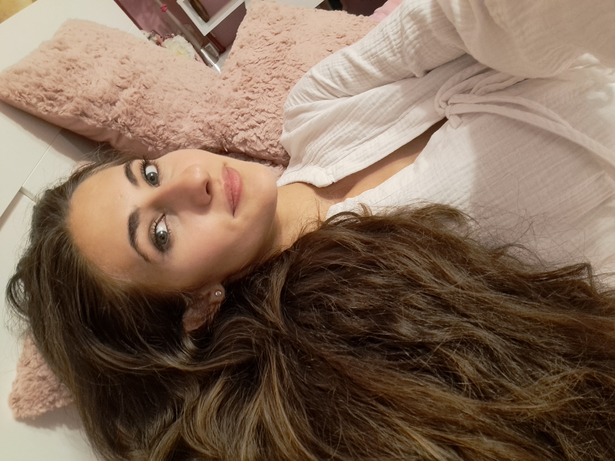 A young adult woman is depicted lying on her back, oriented towards the camera. Her long, wavy, multi-toned brown hair, with visible lighter highlights, is spread out to her left. Her eyes, appearing light green or blue, are open and directed towards the viewer. Her lips are slightly parted in a subtle smile, exhibiting a glossy texture. She wears a small, circular, clear-stoned stud earring in her visible left earlobe. Her attire consists of a white, textured, long-sleeved top with a V-neckline, secured by knotted ties at the chest. She is resting against or beside a fluffy, light pink pillow or cushion. A white surface, consistent with a wall or headboard, is visible behind her to the right. Partially visible to the far right is an indistinct pink object, possibly a box or container. The scene is indoors, in the city of Unknown, Unknown.